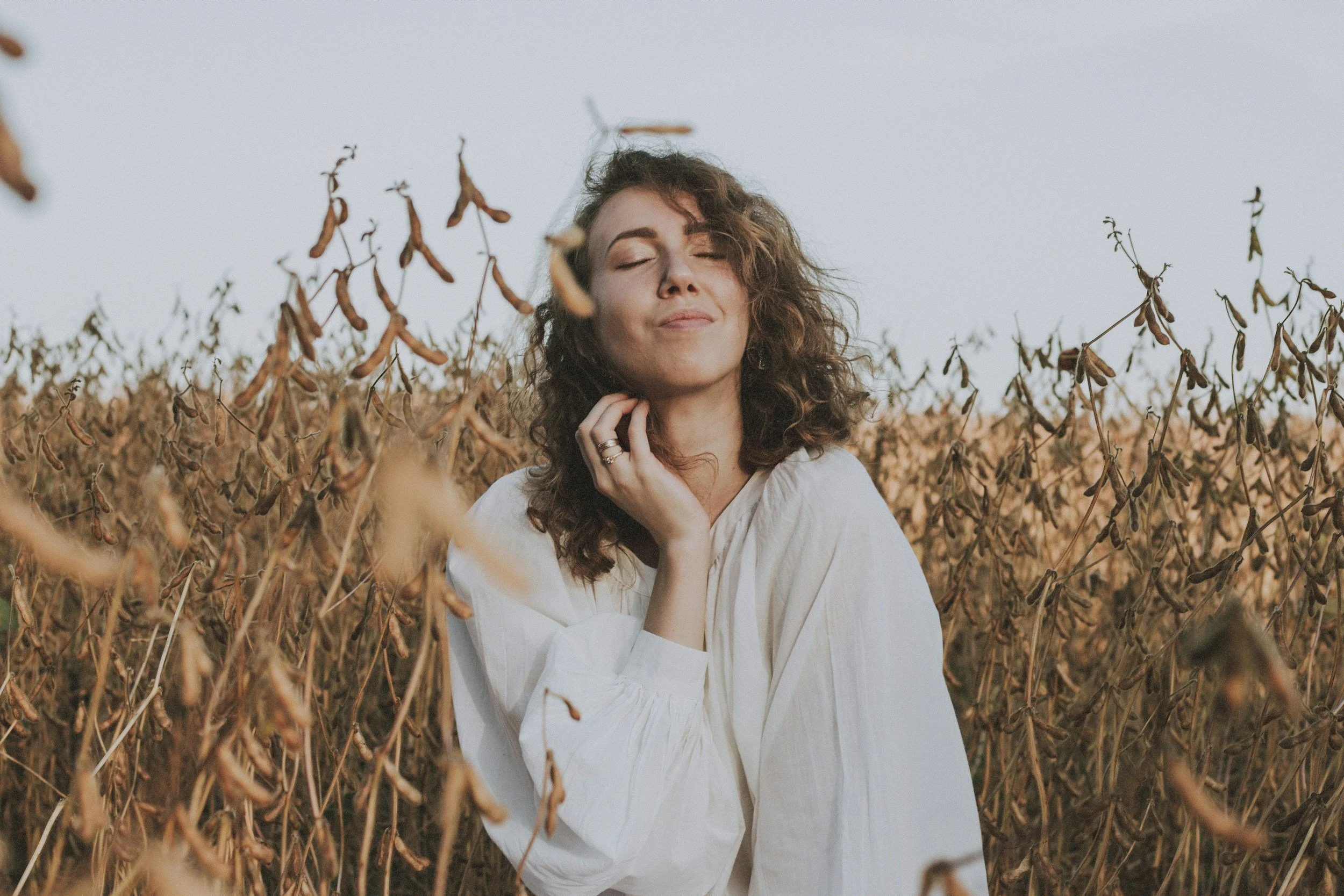A woman with curly hair and closed eyes sitting in a brown field with a clear sky background.