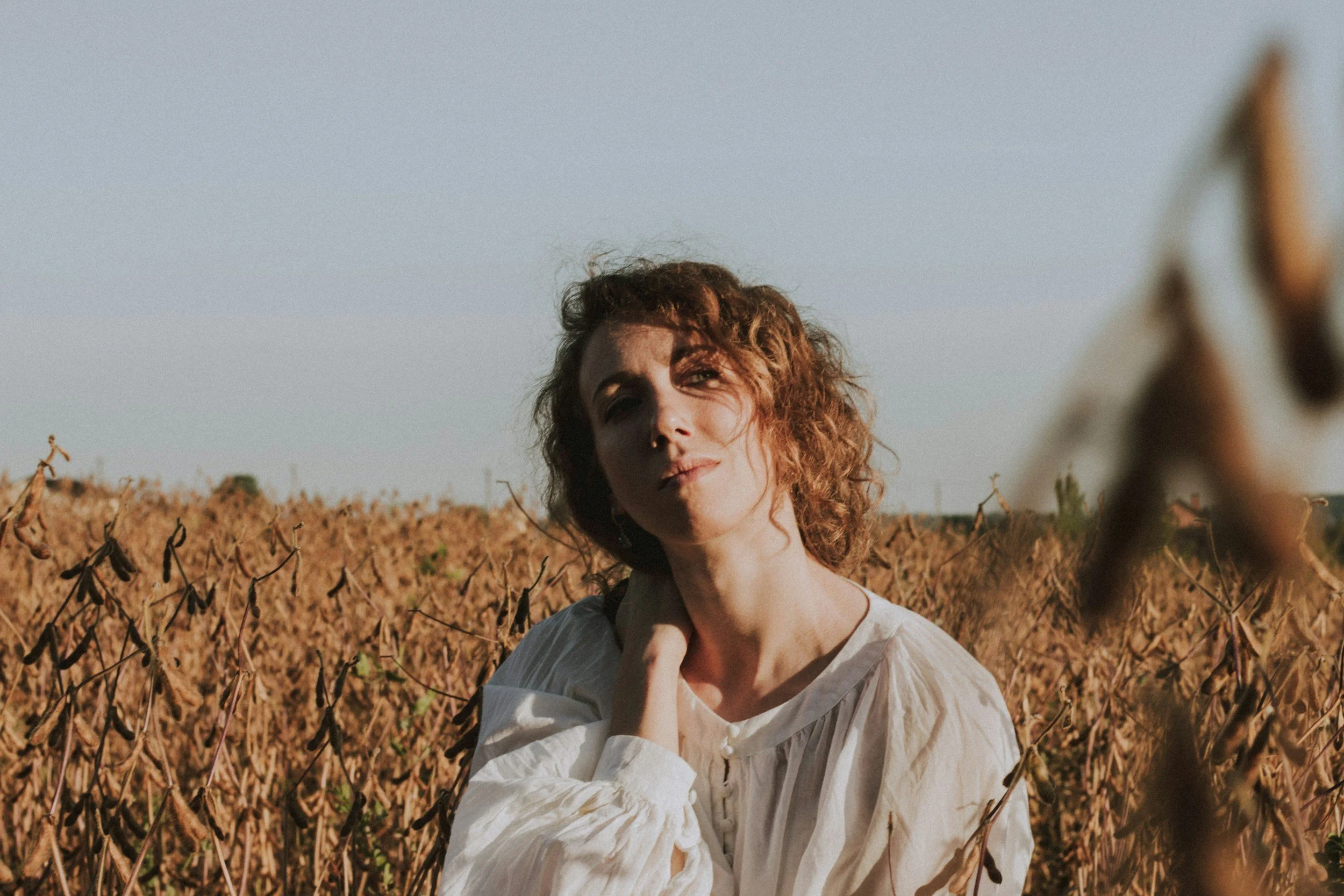 A woman with curly hair standing in a dry, brown field during sunset, wearing a white blouse with one hand near her neck.
