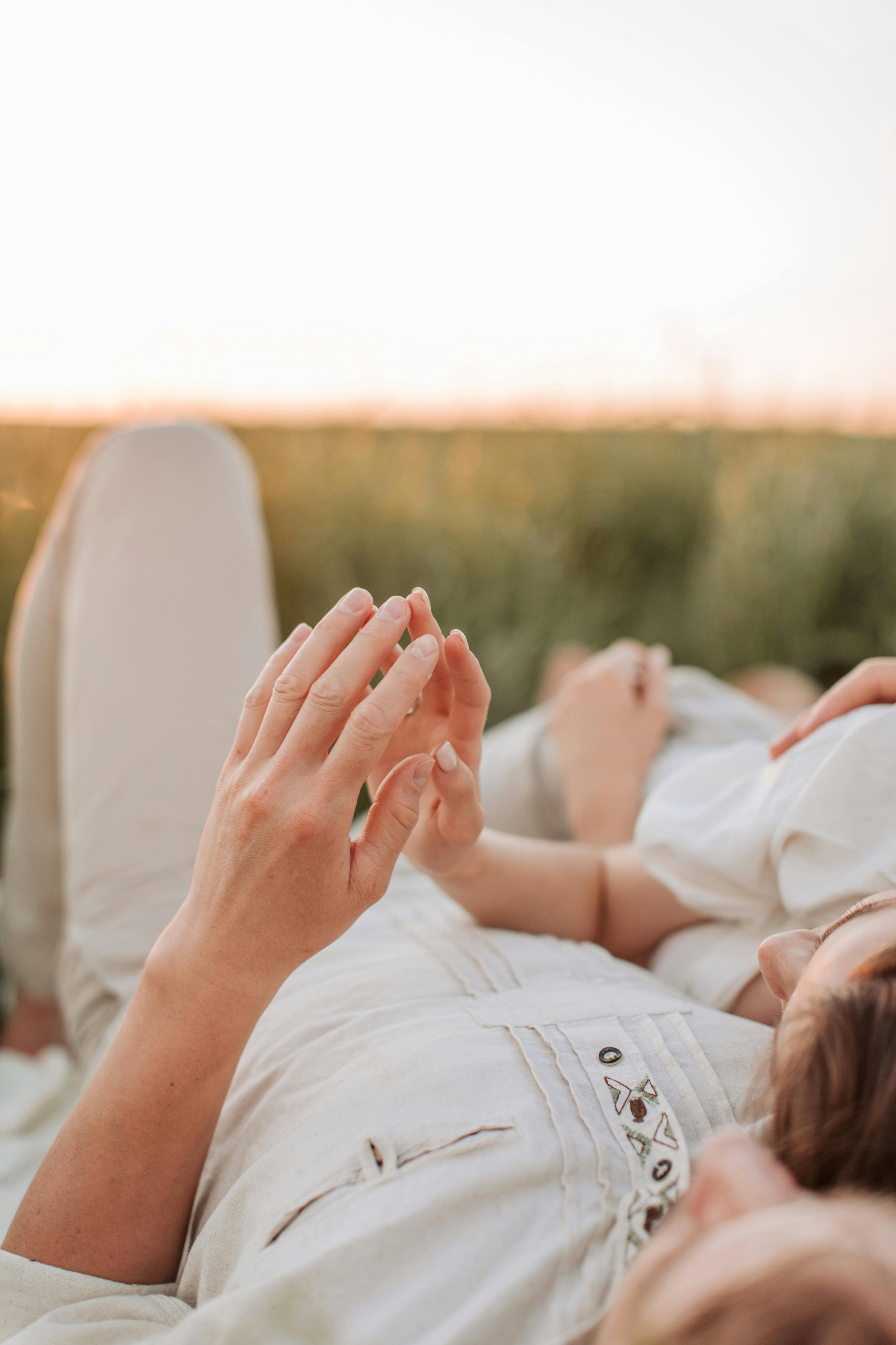 People lying on their backs outdoors, holding hands, with a blurred sunset sky and field in the background.