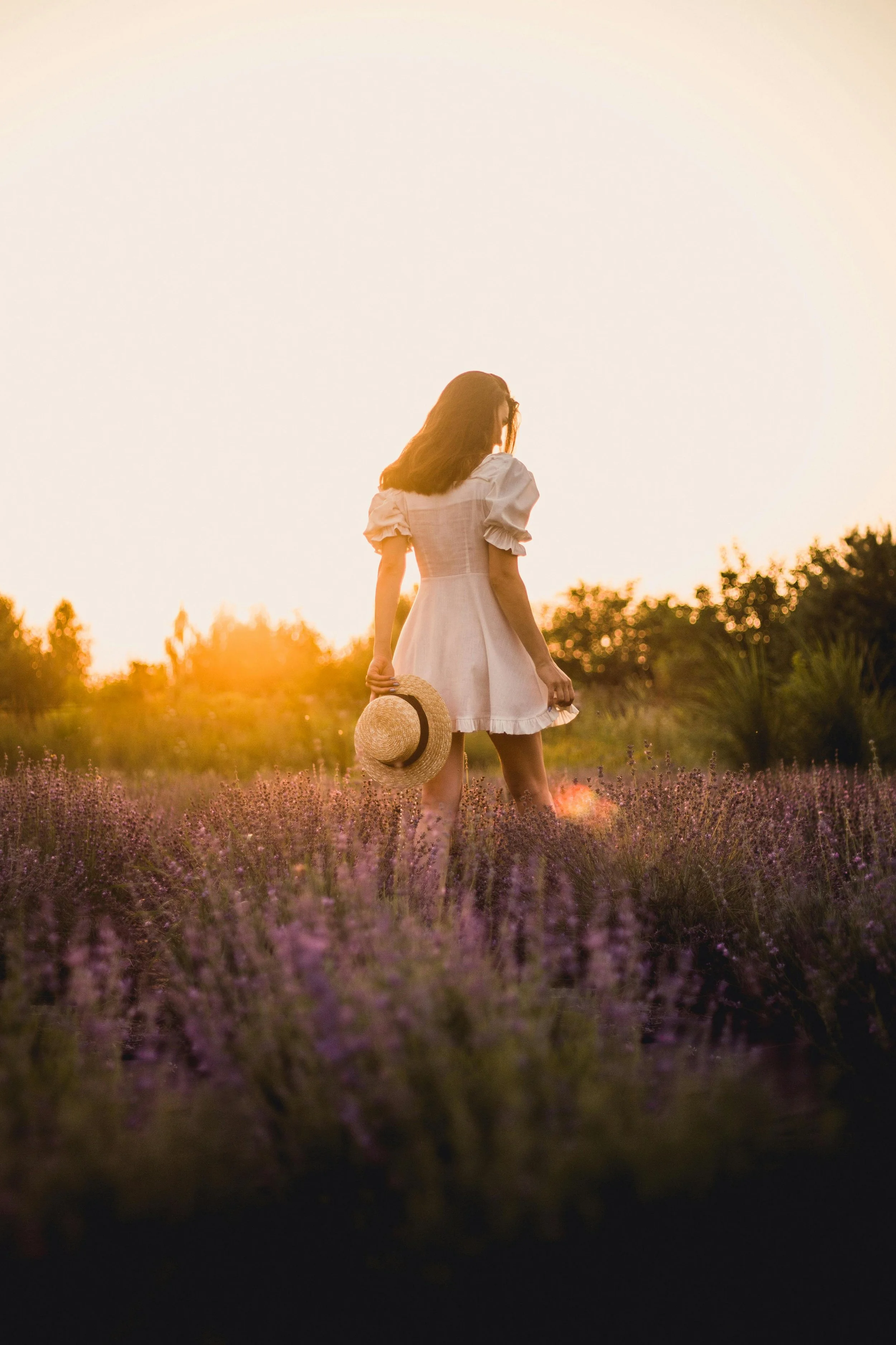 A woman in a white dress holding a straw hat walking through a lavender field at sunset.