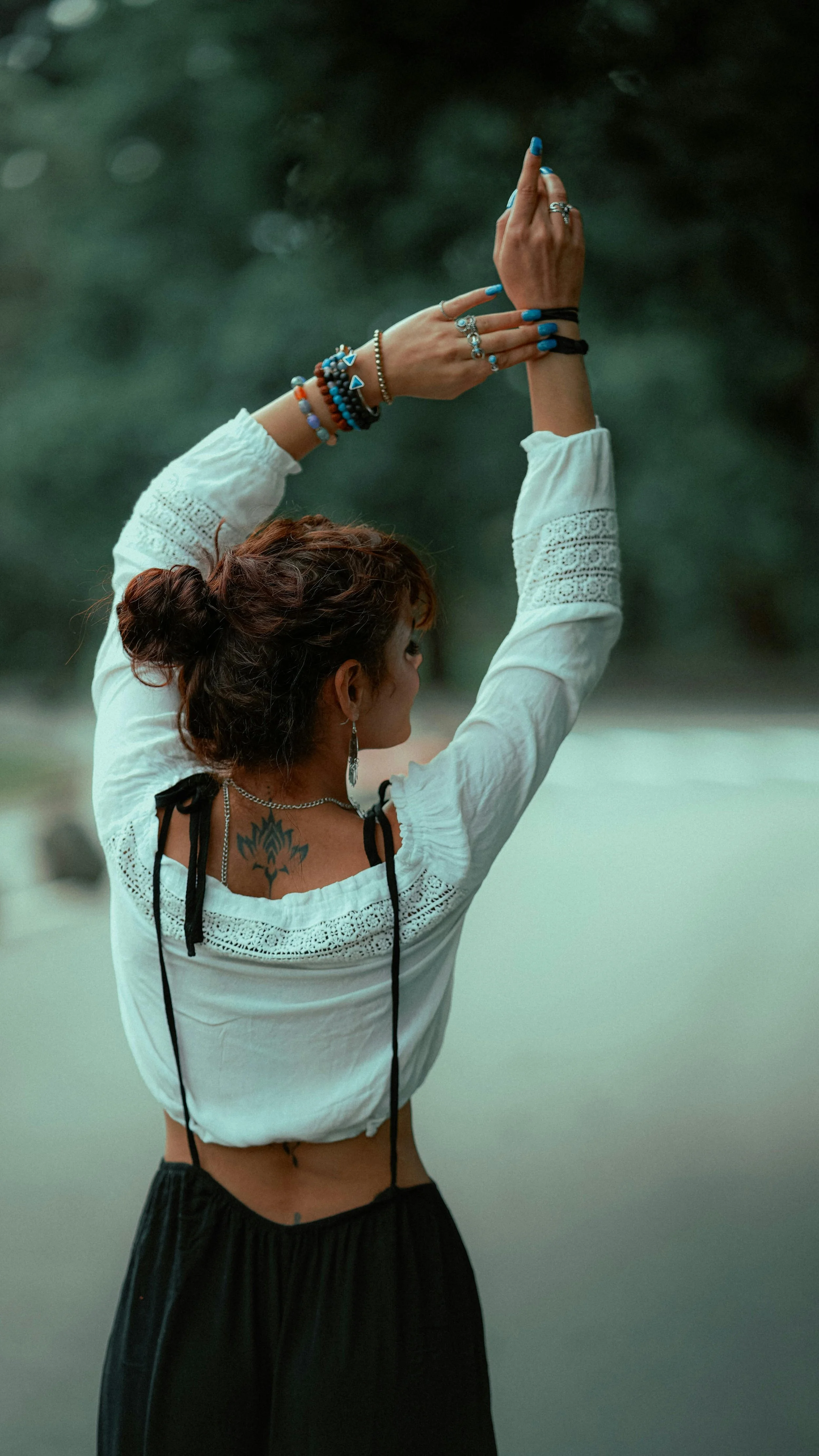 A woman with dark hair in a bun wearing a white embroidered top, black skirt, and multiple bracelets, rings, and jewelry, with tattoos on her back, is raising her hands in a prayer or dance position outdoors during evening.