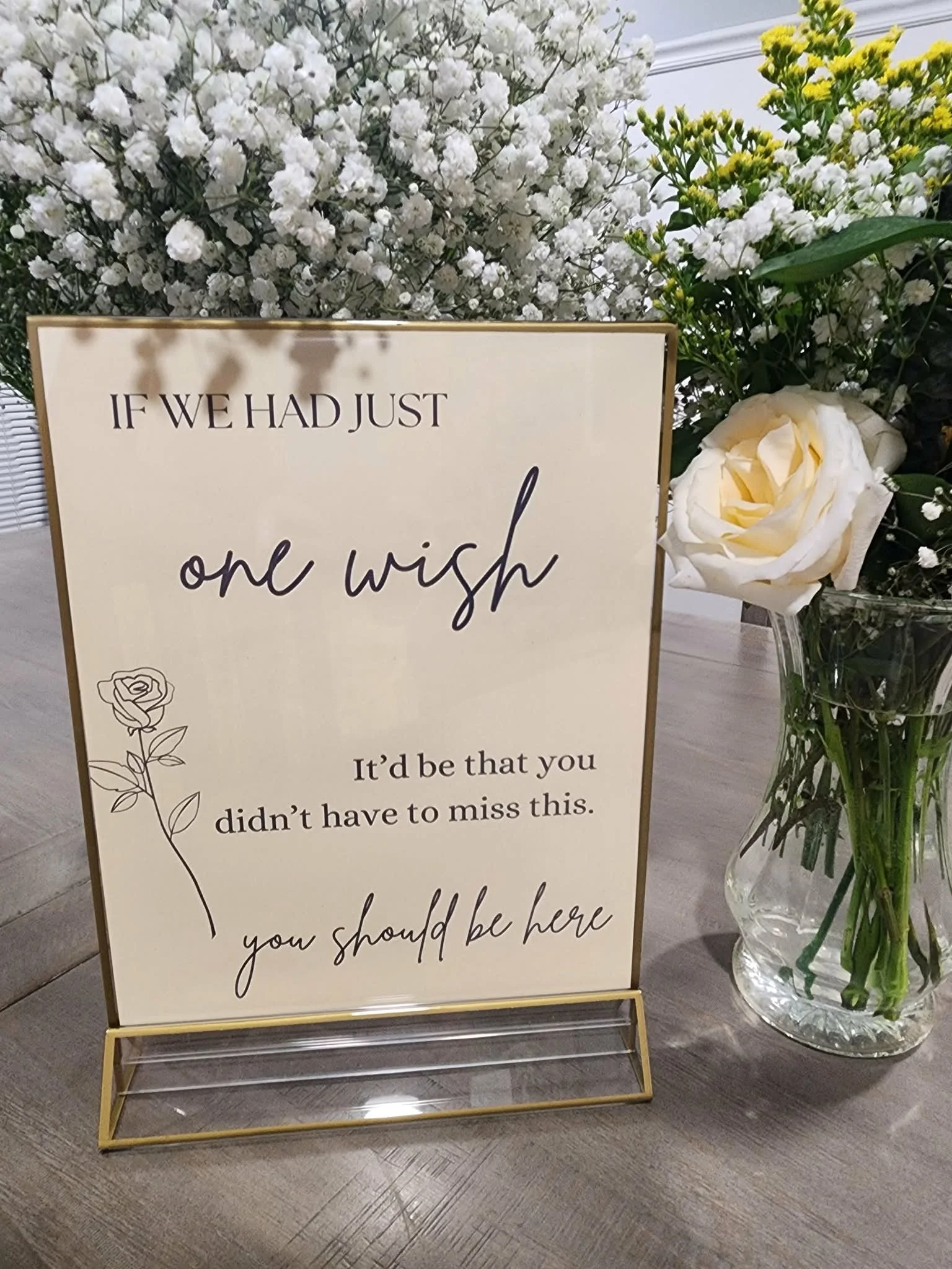 A decorative flower arrangement with white and yellow flowers behind a sign with a heartfelt message. The sign reads: "If we had just one wish, it’d be that you didn’t have to miss this. You should be here." The flowers include white roses and small white and yellow blooms in a glass vase.