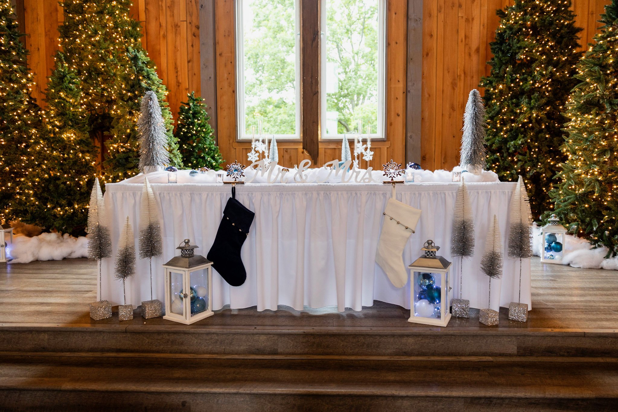A festive holiday display with a table labeled 'Mr. & Mrs.' surrounded by Christmas trees, stockings, lanterns, and ornaments in a wooden room with large windows.