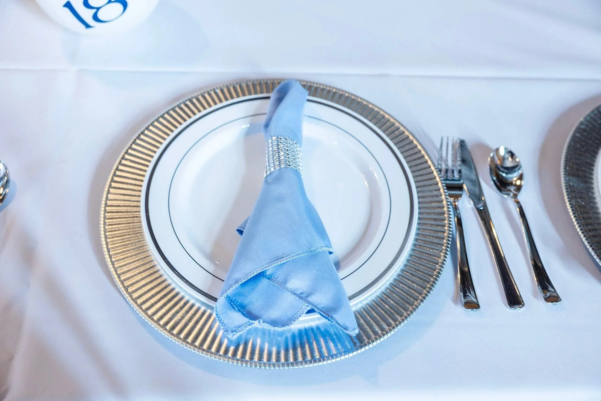 A place setting on a white tablecloth featuring a gold-rimmed plate, a light blue napkin with silver embellishments, a fork, a knife, and a spoon, with a decorative charger beneath the plate.