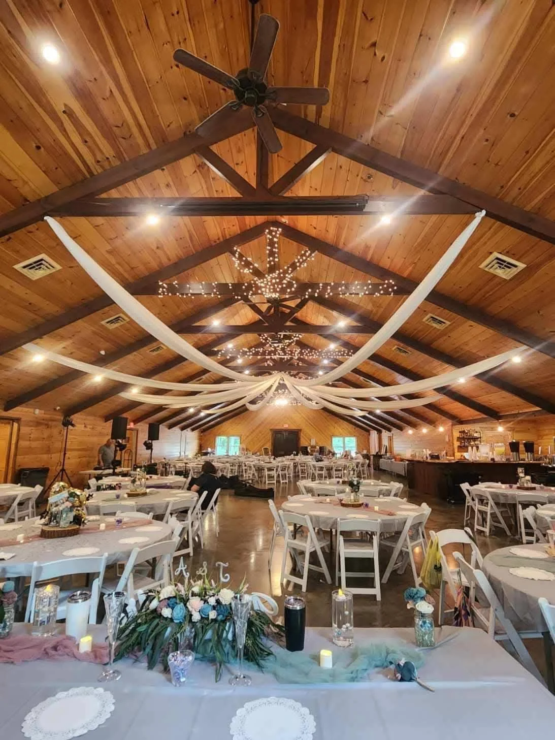 Indoor event space decorated with fairy lights, draped fabric, round tables with floral centerpieces, and a wooden ceiling with ceiling fans.