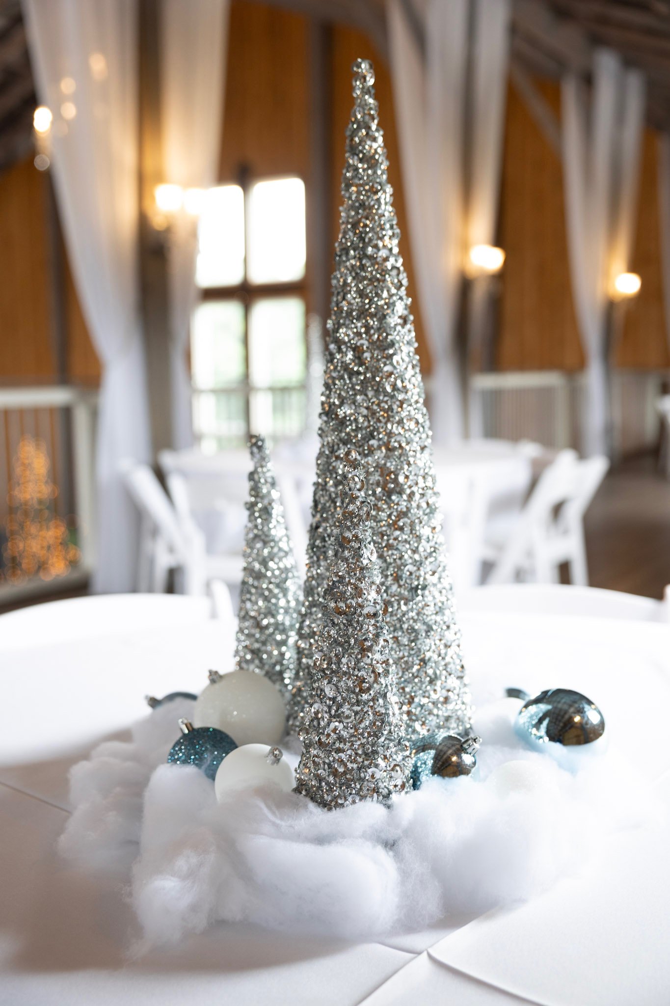 Silver glittery cone-shaped decorations resembling Christmas trees on a white cotton-like surface, surrounded by blue, silver, and white baubles, with a background of a wooden interior with large windows and white curtains.
