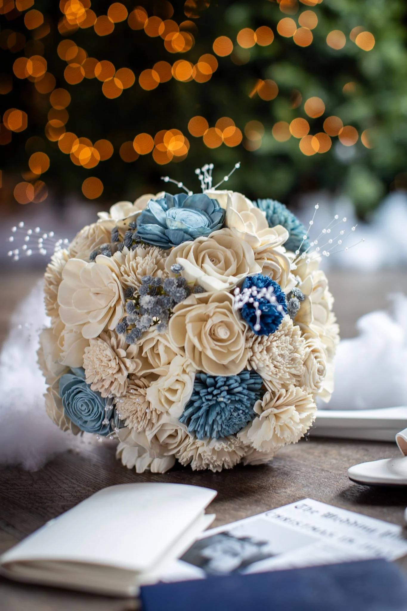 A bouquet of cream and blue paper flowers on a table, with a blurred background of warm bokeh lights.