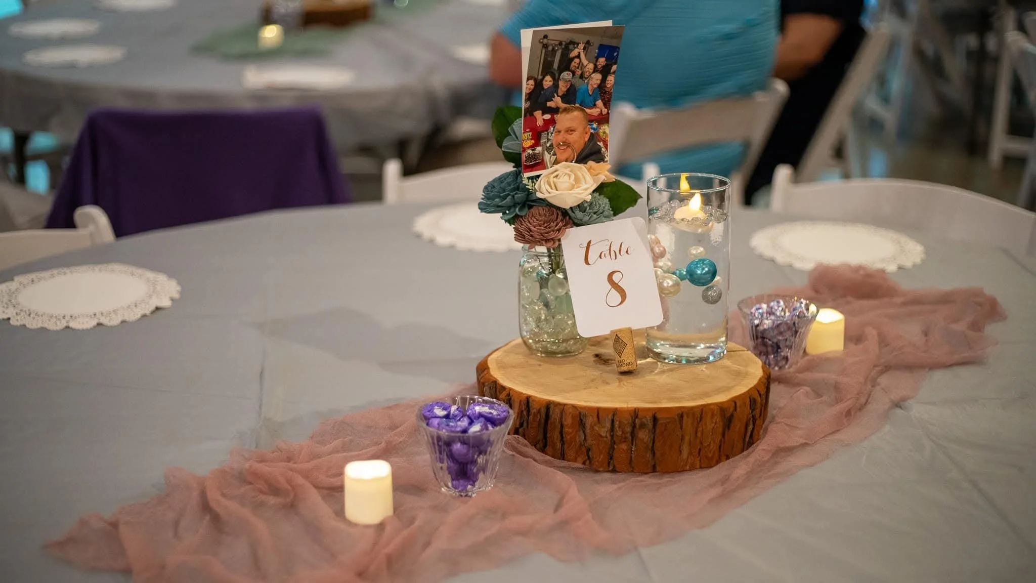 Wedding reception table centerpiece featuring a small glass jar with flowers, a table number sign, candles, and decorative beads on a wooden slab, with a pink fabric runner underneath.