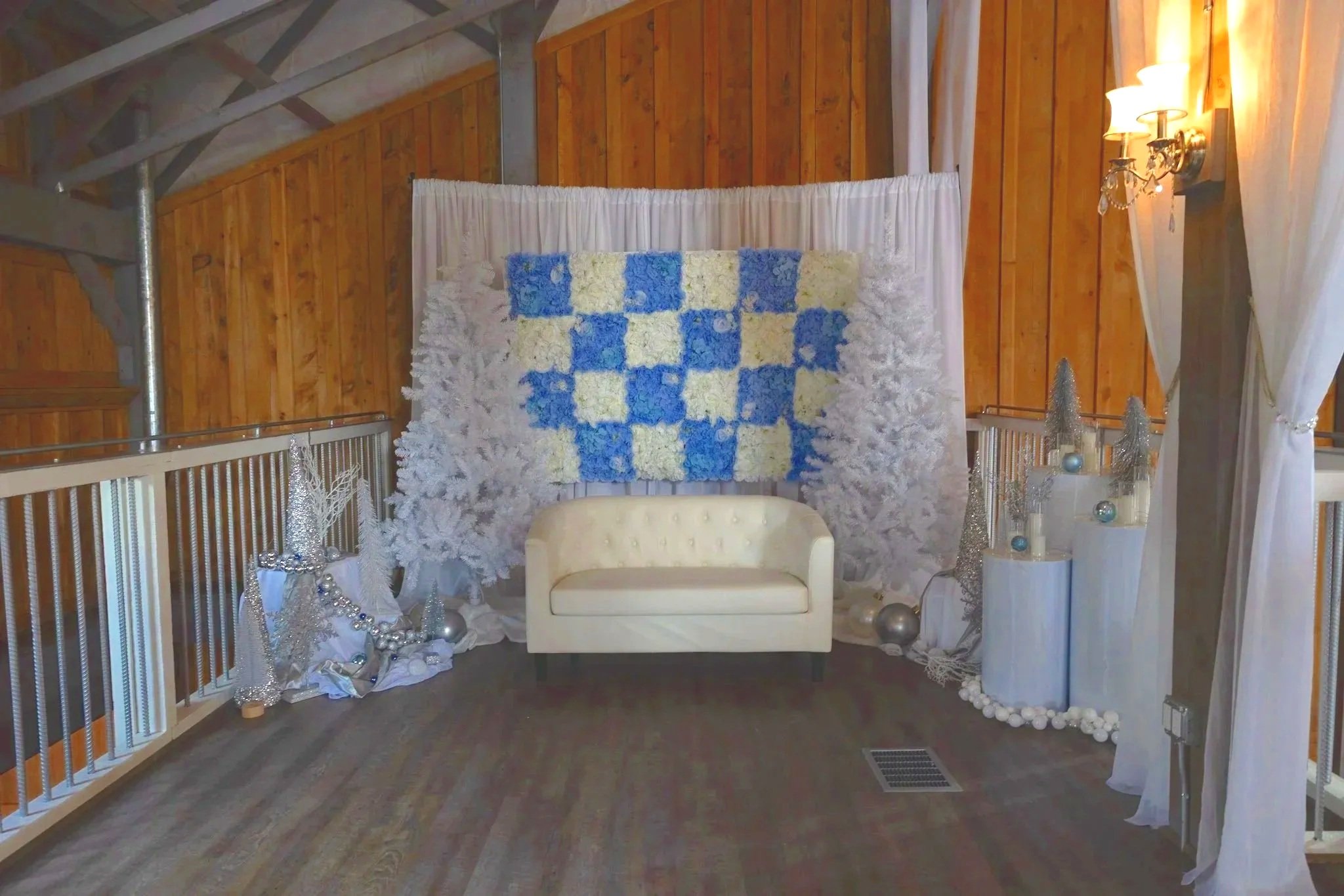 Indoor space decorated for winter or holiday season, with a white and blue floral tapestry hanging on a wooden wall, white curtains, a white loveseat, silver and white holiday ornaments, and decorative trees.