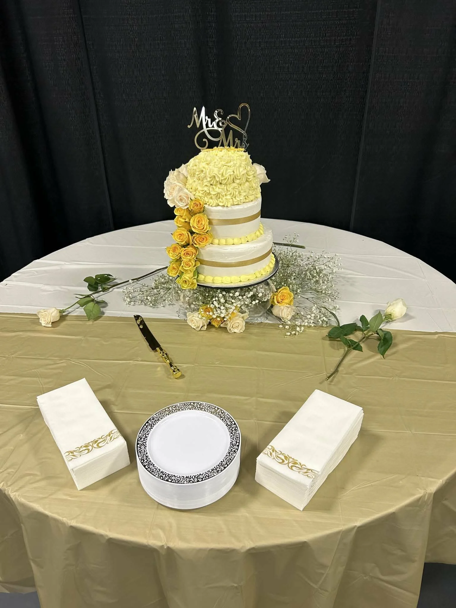 A wedding cake with two tiers, decorated with yellow roses, white roses, and baby's breath, topped with a cake topper reading 'Mr. & Mrs.' on a table covered with a gold cloth, surrounded by white napkins, a knife, and paper plates with gold and black patterns. The background is black.