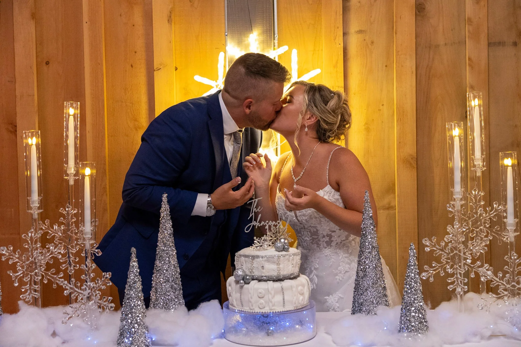 A bride and groom sharing a kiss at their wedding cake with gold and silver decorations behind them.
