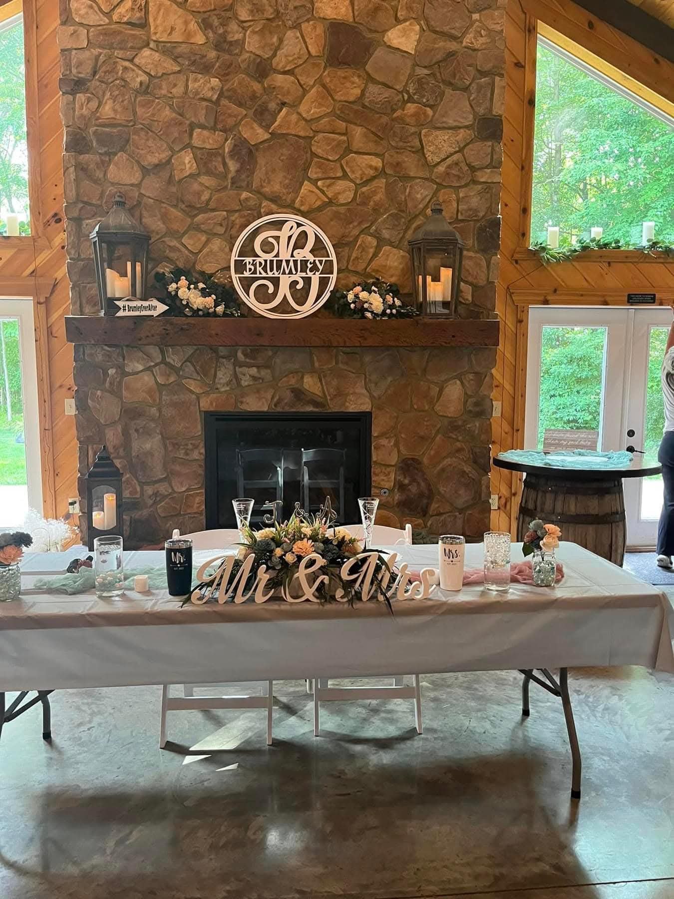 Decorations for a wedding reception in a rustic setting. A table with a cake topper reading 'Mr & Mrs' and floral arrangements. A fireplace with 'The Brumley' sign, lanterns, and candles. Large windows revealing green trees outside.