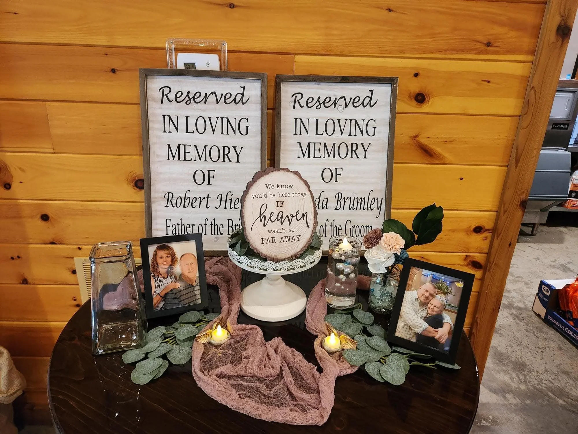 A memorial table with framed photographs of a couple, candles, and a sign that reads 'Reserved in loving memory of Robert Hie' and 'In loving memory of Linda Brumley.' The table is decorated with greenery and pink fabric, all set against a wooden wall.