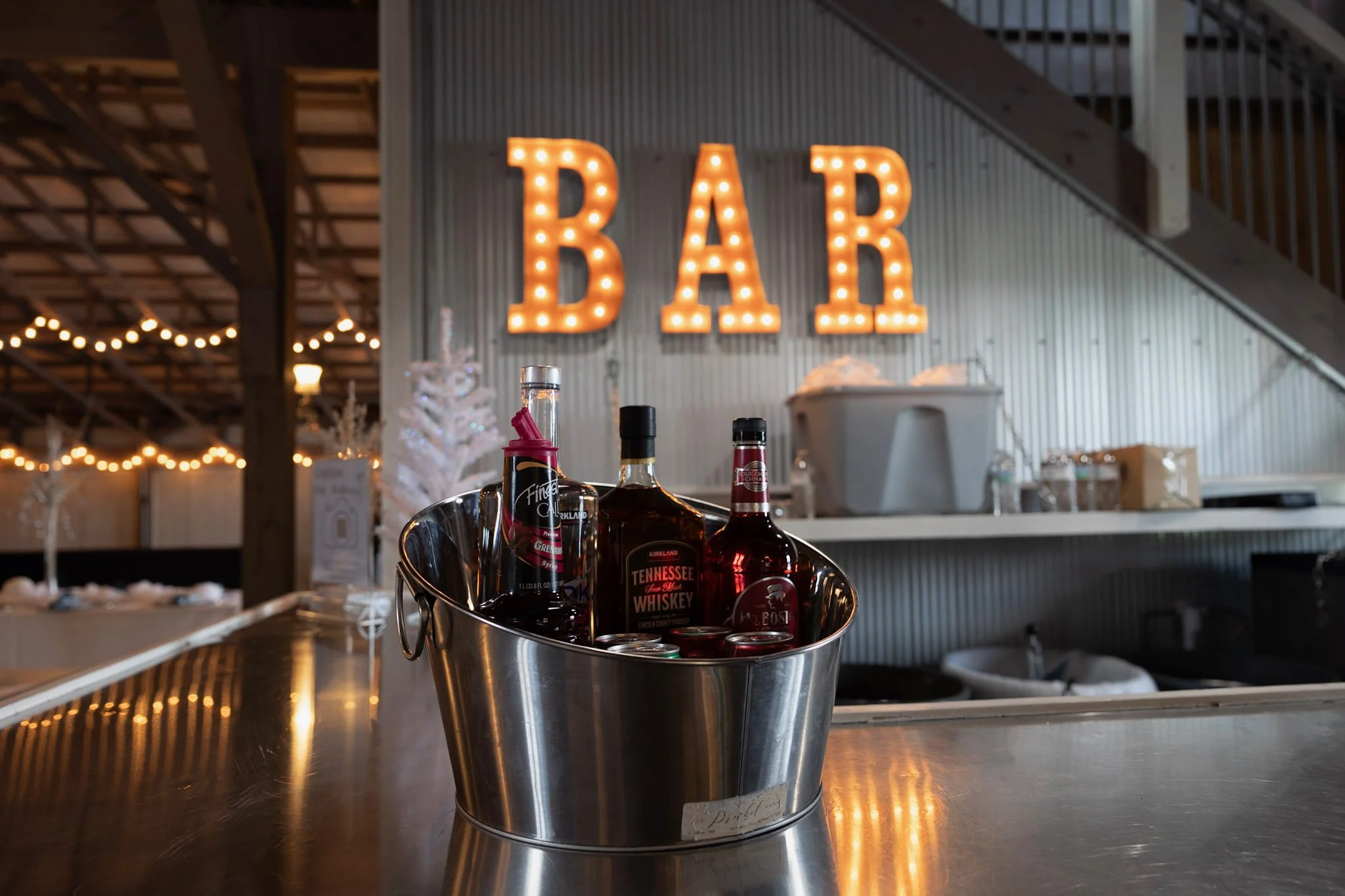 A metal bucket with bottles and cans of whiskey and soda on a bar counter in front of a lit 'BAR' sign with warm lighting and a rustic interior.