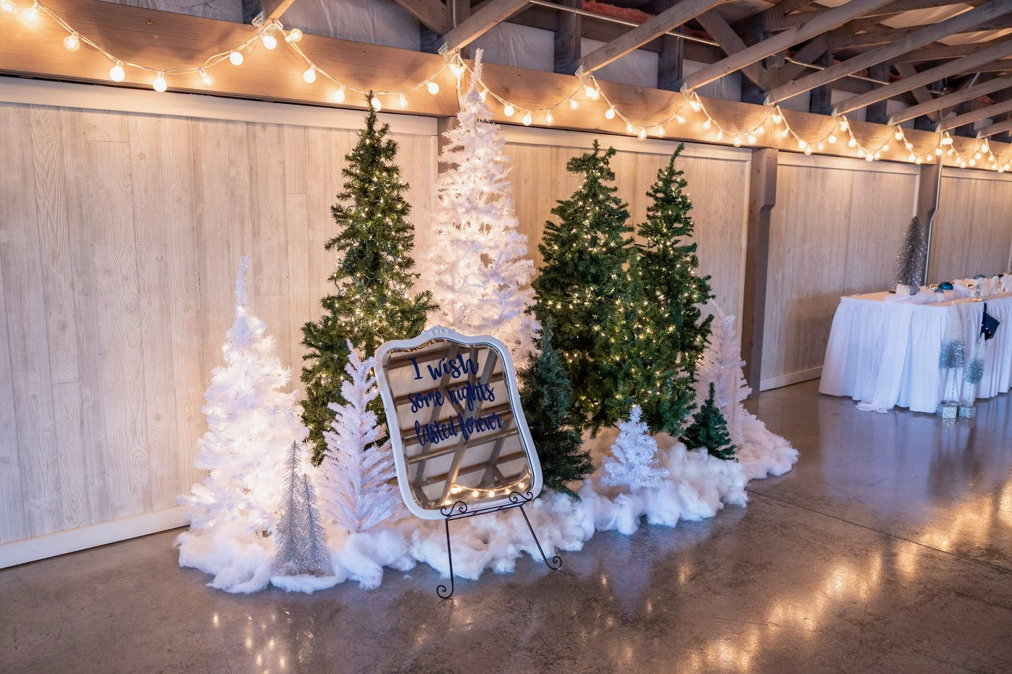Decorative Christmas setup with artificial snow-covered white and green Christmas trees with string lights hanging overhead, a sign that reads 'I wish some nights lasted forever,' and a table with holiday decorations on the right side.