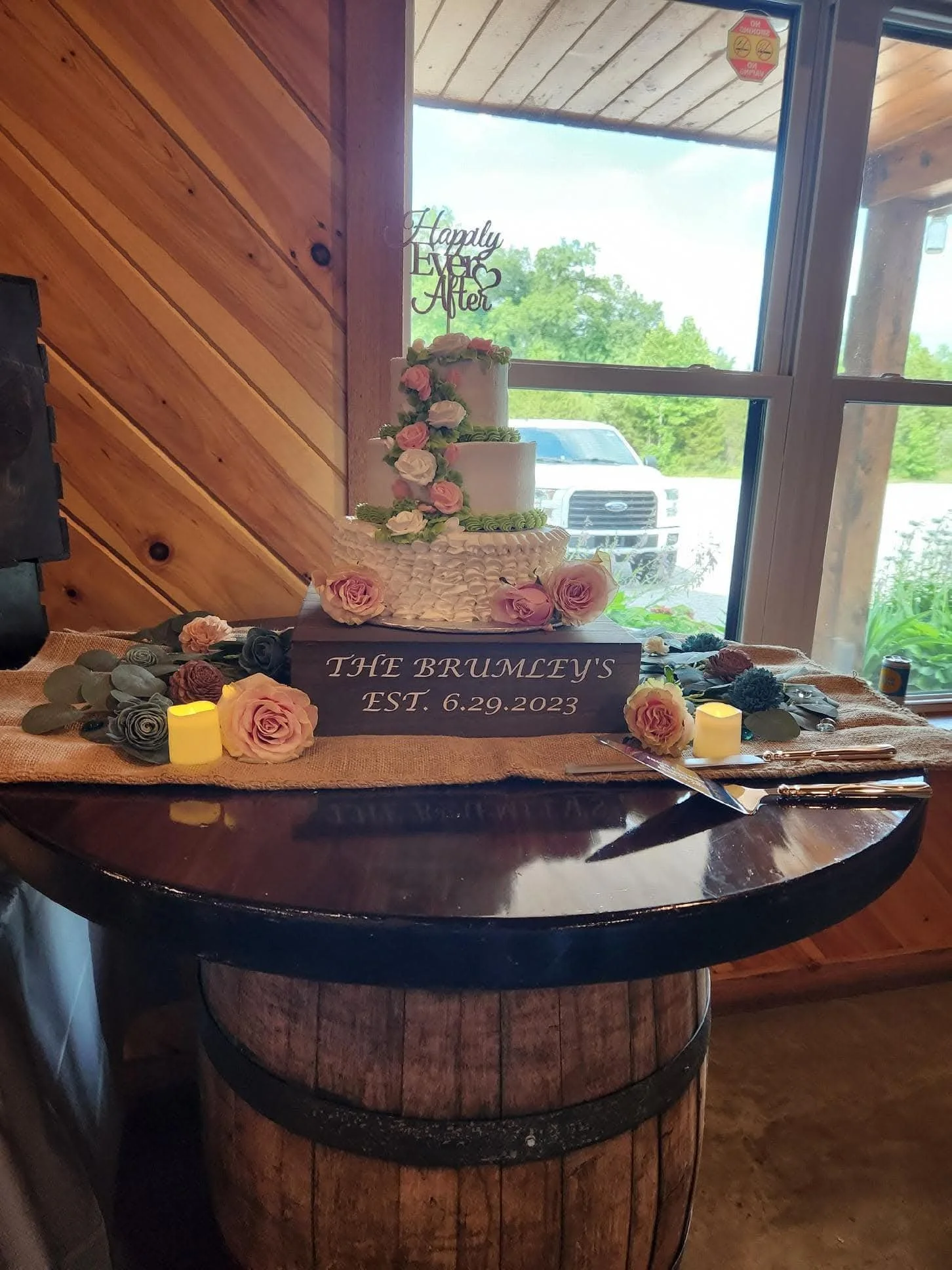 A wedding cake with pink and white flowers on a wooden barrel table, decorated with a sign that reads 'The Brumley's Est. 6.29.2023' and a cake topper that says 'Happily Ever After.'