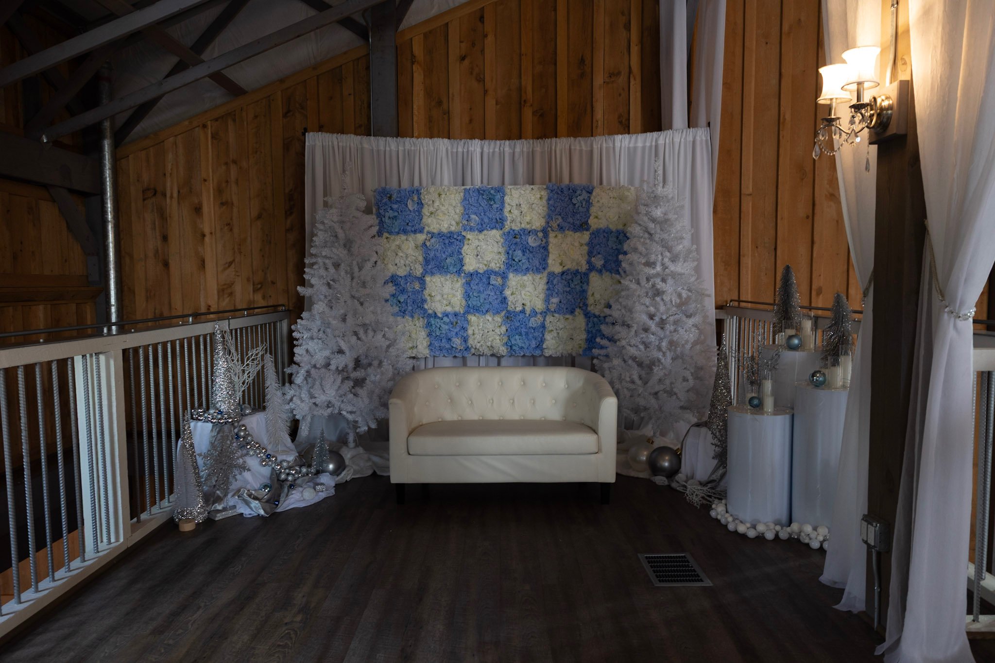 A cozy indoor space decorated for a winter or holiday event with a white and blue floral quilt hanging on a wall, white Christmas trees, silver ornaments, and white drapery.