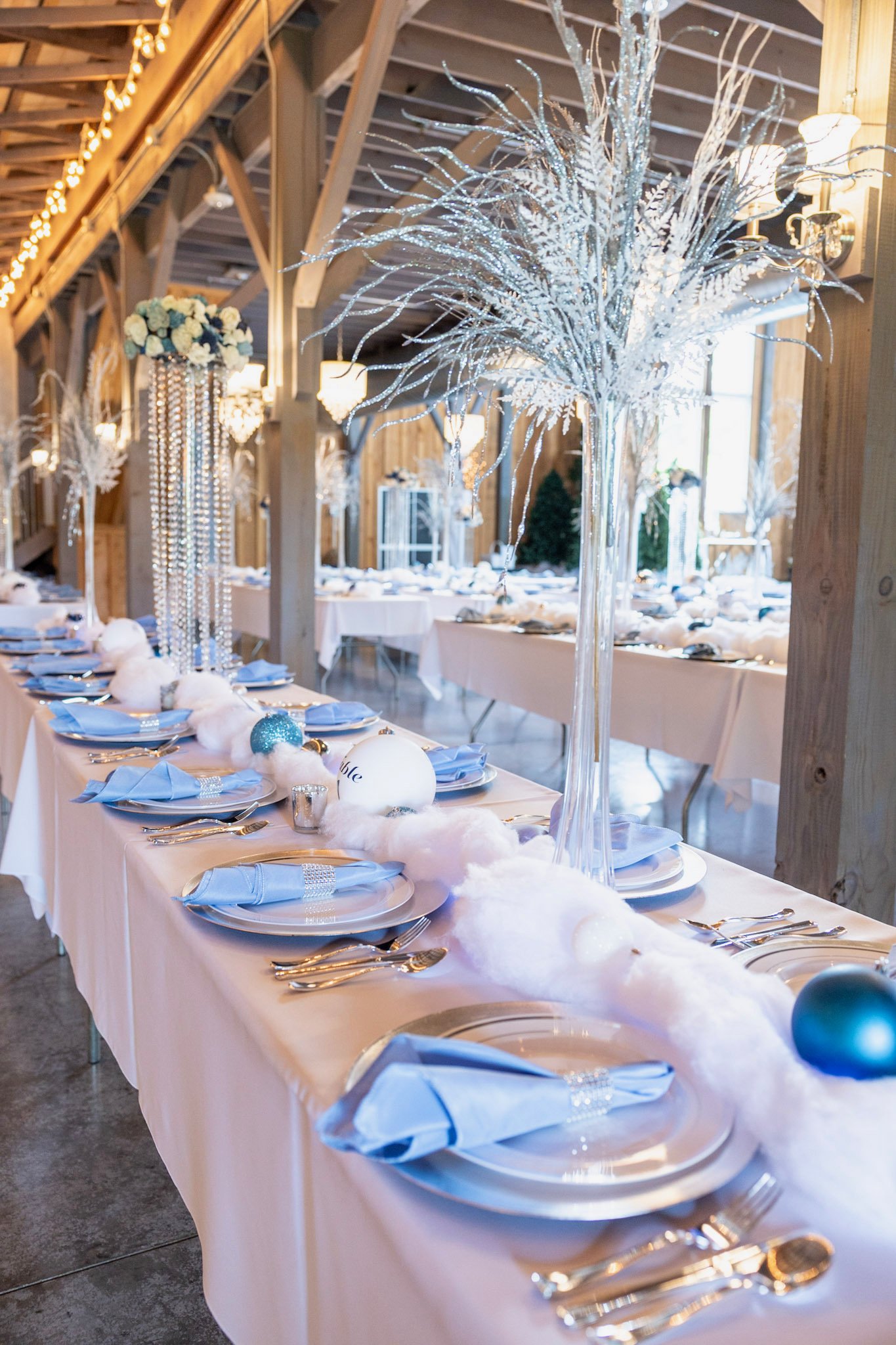 A decorated banquet table set for a celebration with white tablecloths, blue napkins, and silverware, featuring tall centerpiece arrangements with white and silver ornaments and ribbons inside clear vases, illuminated in a wooden barn-style venue with warm lighting.