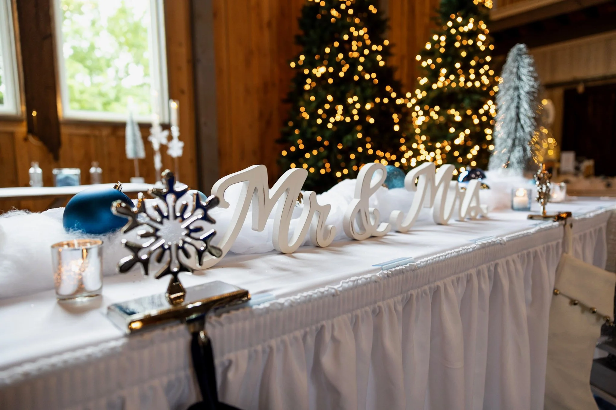 A decorated table with a white tablecloth, illuminated candles, snowflake decorations, and a large white 'Mr & Mrs' sign. In the background, a lit Christmas tree and a window with a view of green trees.