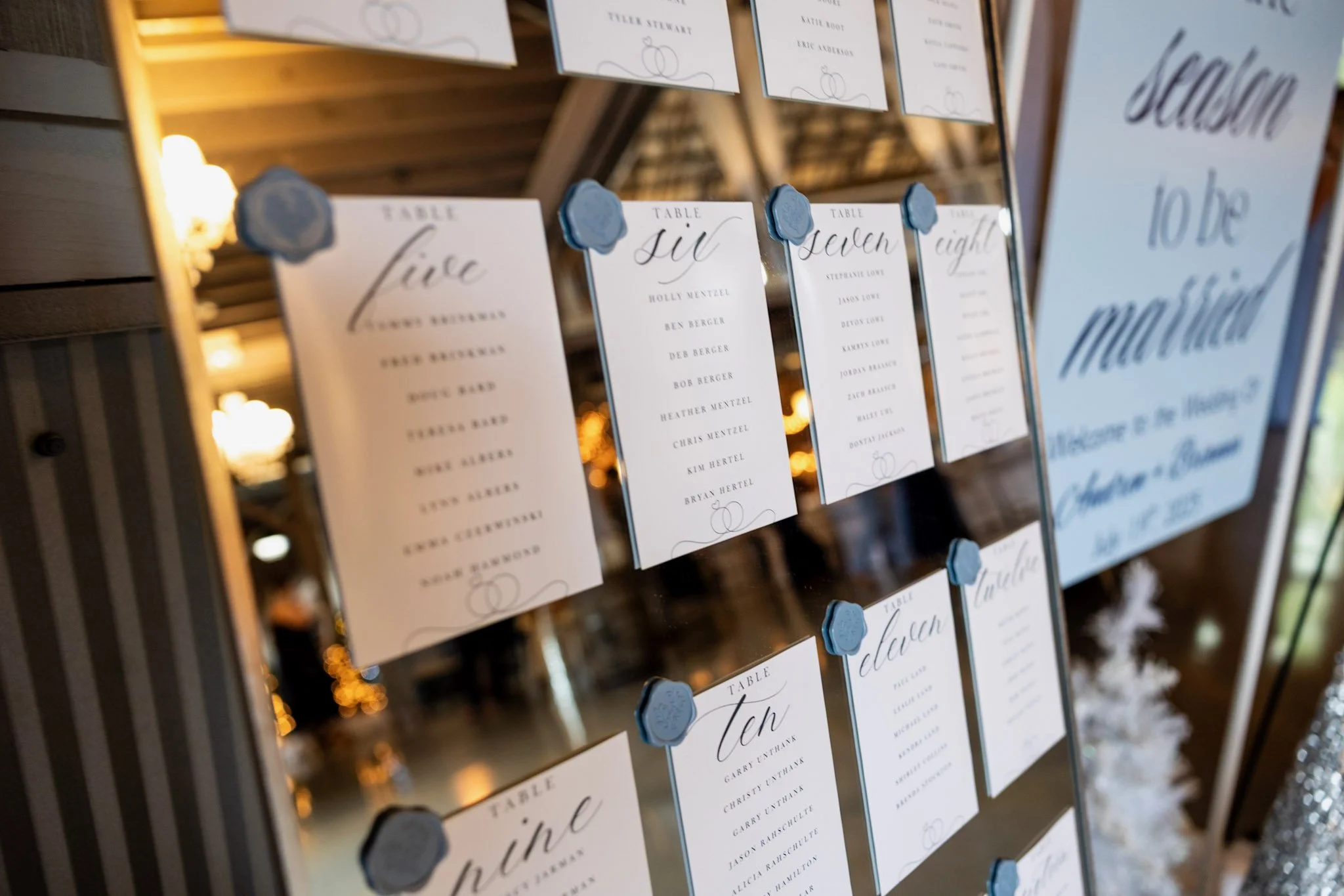 Wedding seating chart with handwritten-style cards listing guest names under table numbers, attached with blue wax seal stickers, displayed on a mirror.