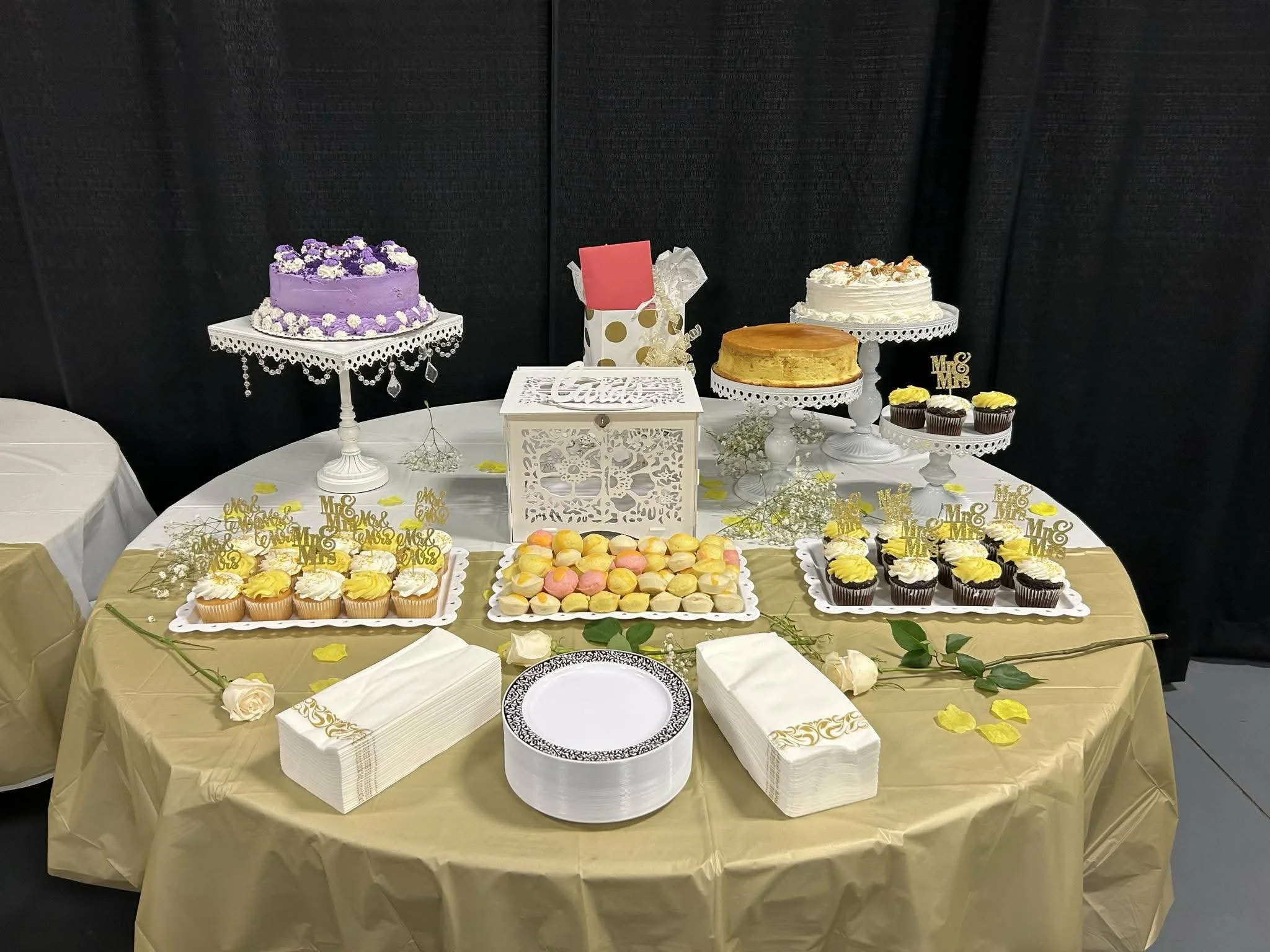 A dessert table with various cakes, cupcakes, and sweets, decorated with yellow and white accents and 'Mr. & Mrs.' signs, set against a black backdrop.