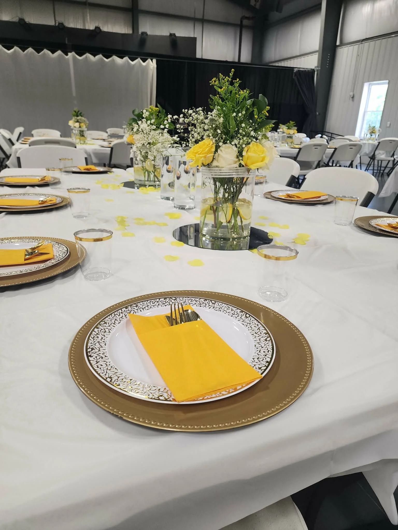 A decorated banquet table set with gold and white dinnerware, yellow paper napkins, and a floral centerpiece with white and yellow roses and baby's breath in a glass vase, in a large hall with industrial walls and high ceilings.