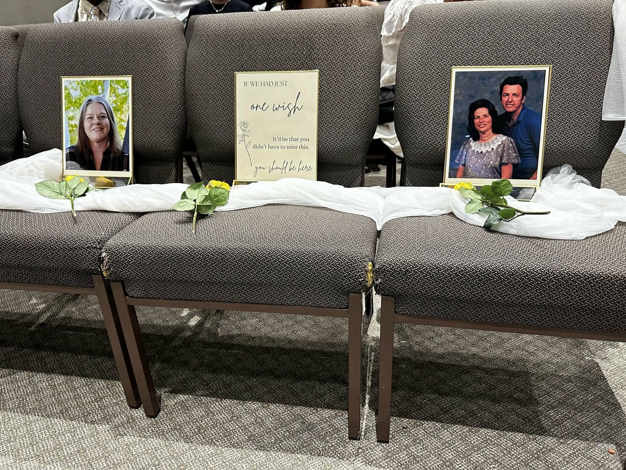Memorial display on chairs with photographs of a woman and a man, a framed message sign, and floral decorations at a ceremony.