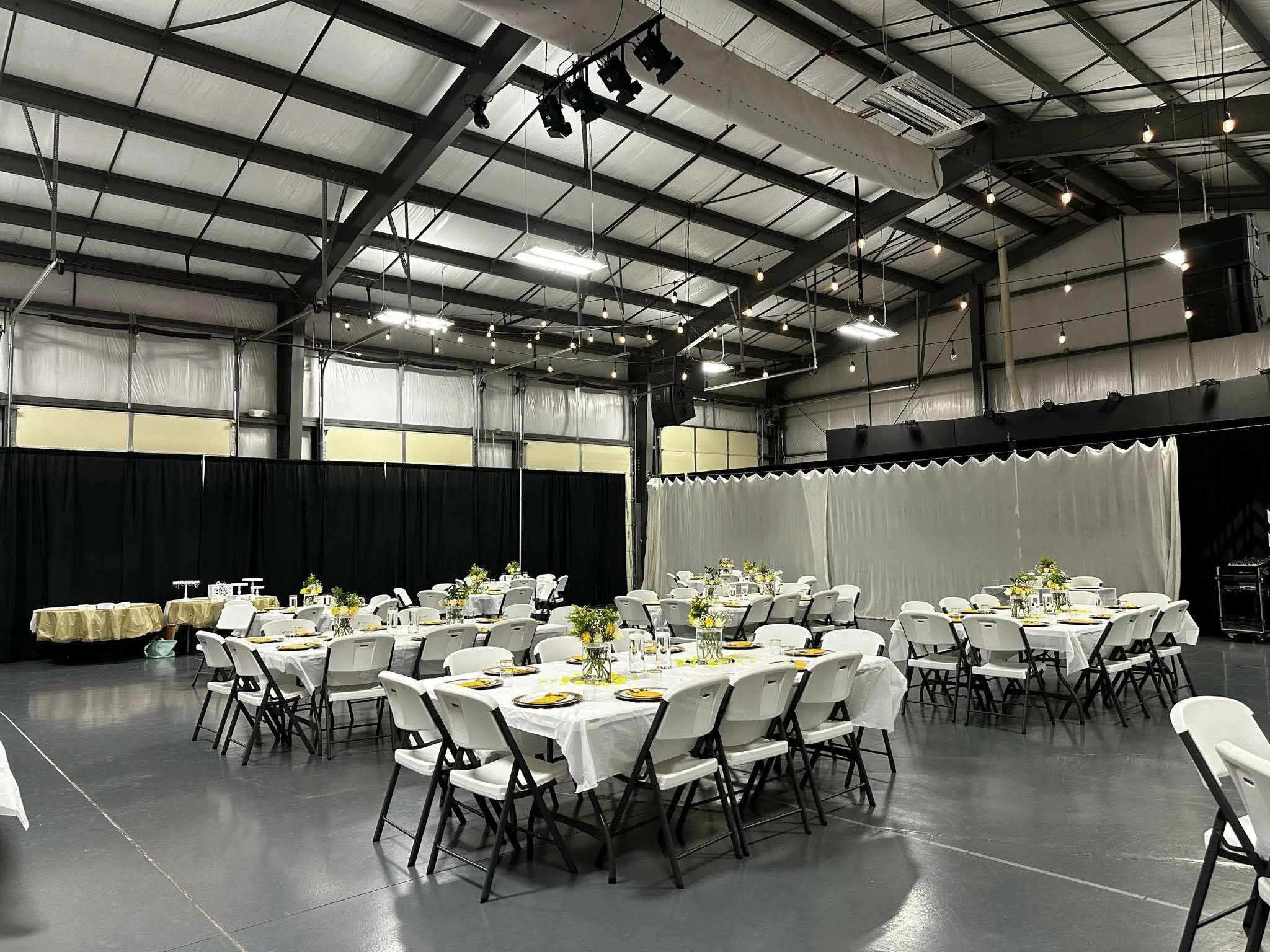 Indoor event space with round tables covered in white tablecloths, decorated with yellow flowers in glass vases, and surrounded by white chairs. The ceiling is high with black metal beams and hanging string lights. There are black curtains along the walls and some stage or service equipment at the back.