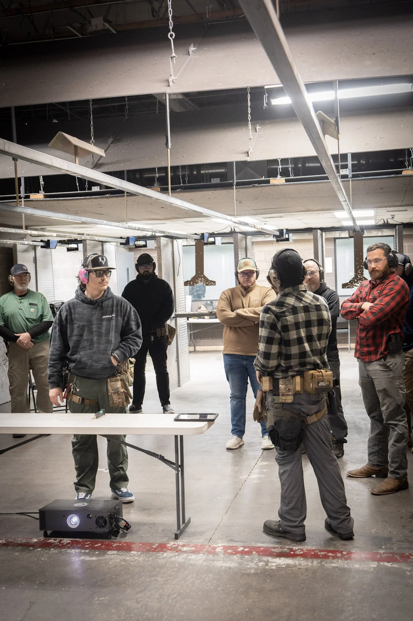 A group of men, wearing safety headphones and tactical gear, are gathered around at an indoor Tactical Penguin range. They are standing near a table with target equipment and a projector on the floor, with an industrial ceiling above.