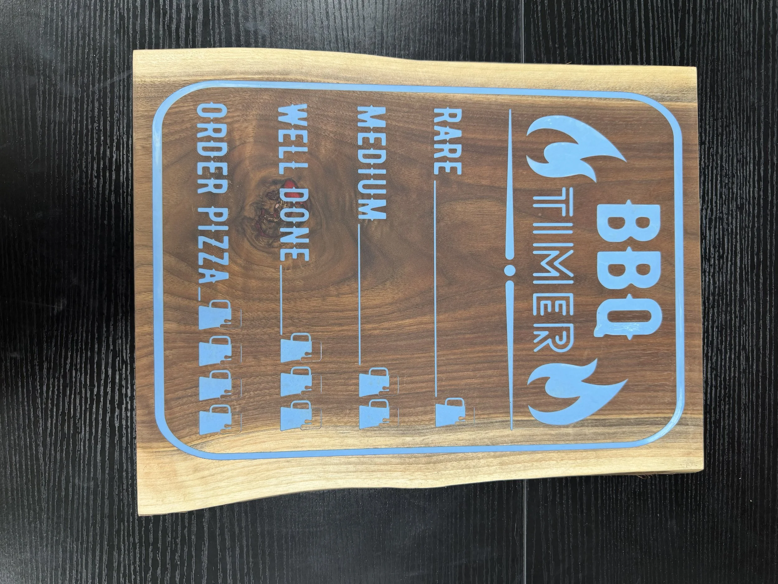 A wooden rectangular sign engraved with blue text and icons. The sign has a flame logo, the message "BBQ Timer" and a list of instructions about cooking times.
