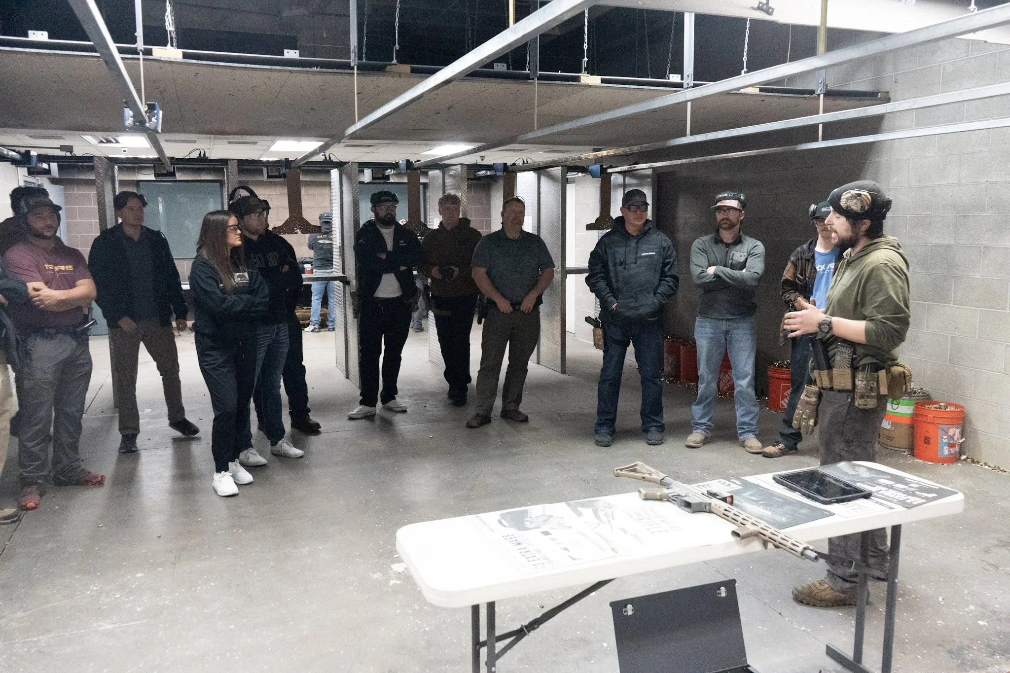 Group of people standing and listening to a person in tactical gear in an industrial or workshop setting with tools and equipment on tables.
