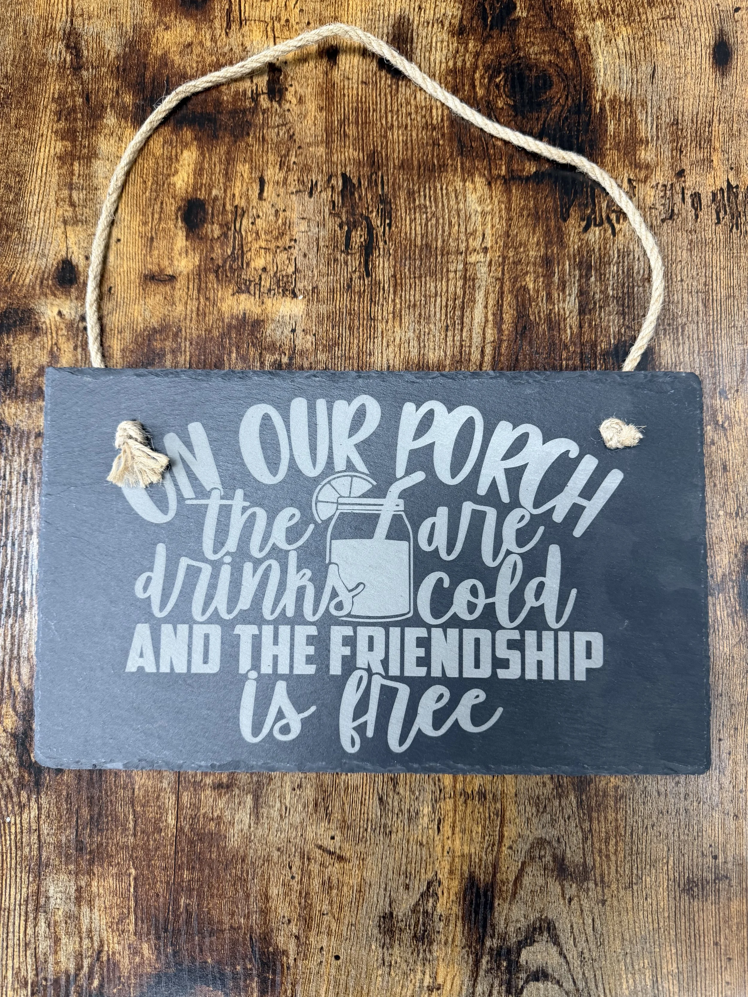 A black chalkboard sign with white text that reads, 'On our porch the drinks are cold and the friendship is free,' hung by a piece of twine on a wooden surface.