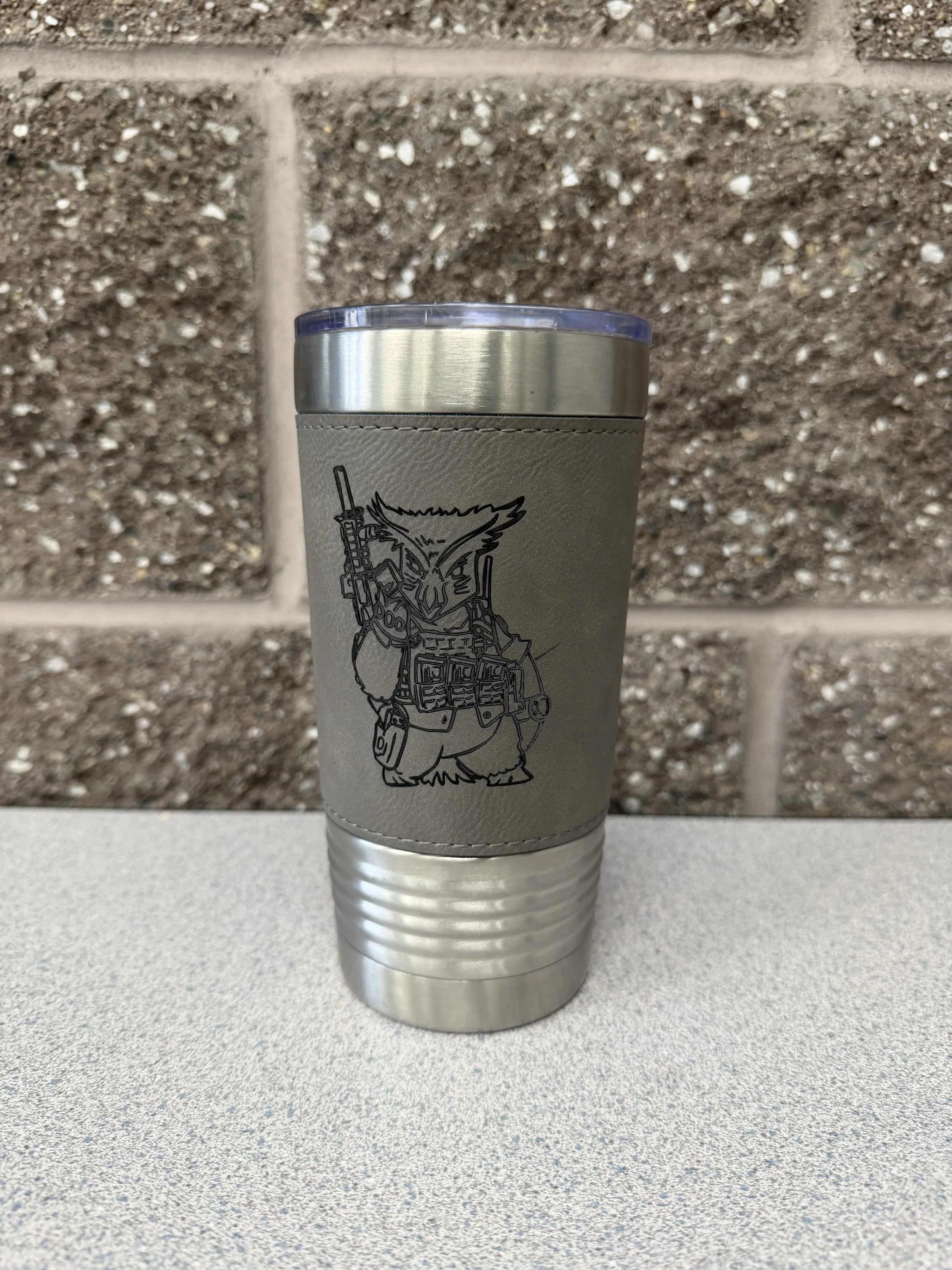 A cylindrical silver tumbler with a gray leather sleeve featuring an engraving of the Tactical Penguin logo.