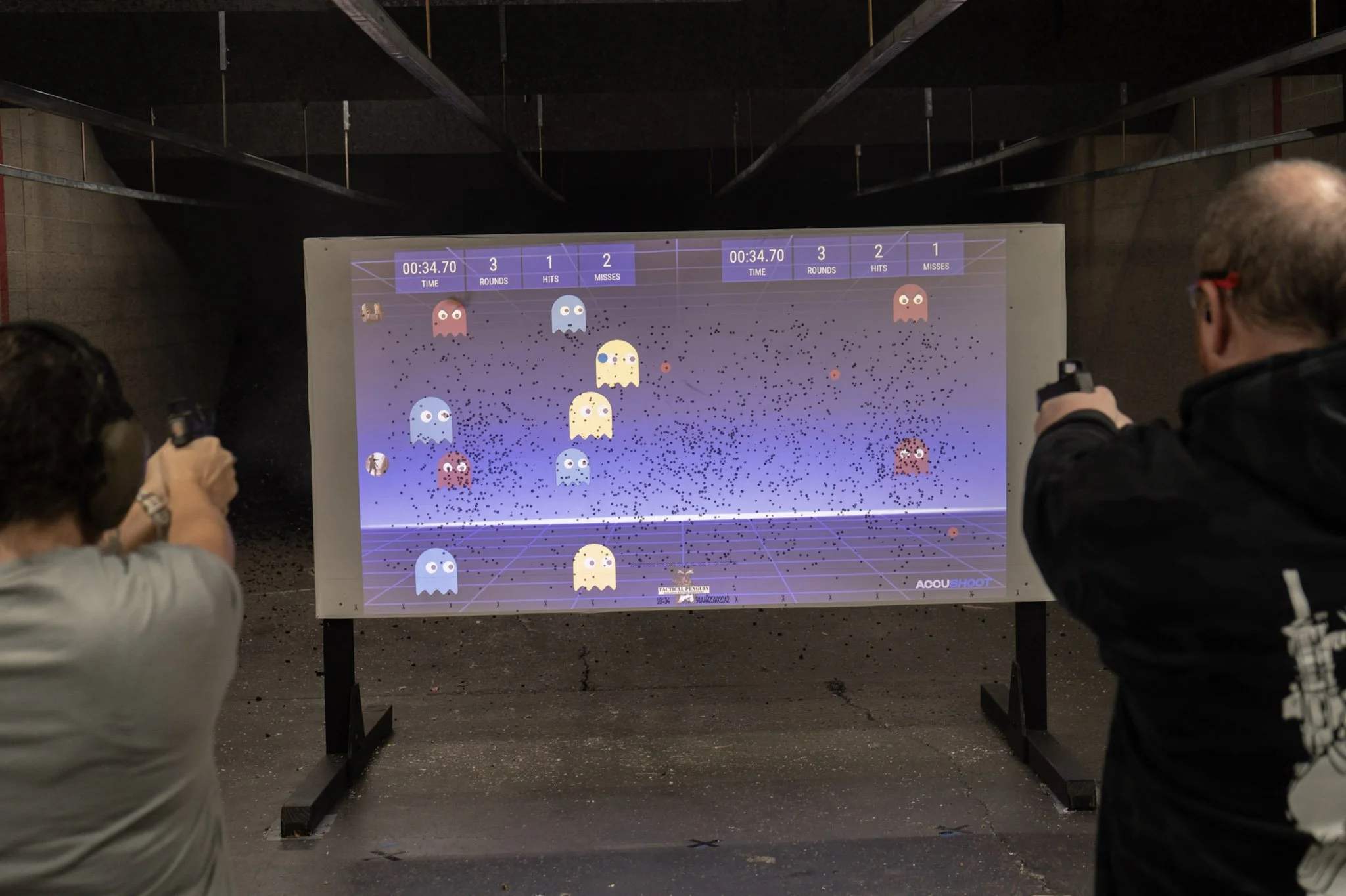 People playing a shooting arcade game on a large screen in an indoor shooting range, with guns aimed at the screen featuring cartoon ghosts.