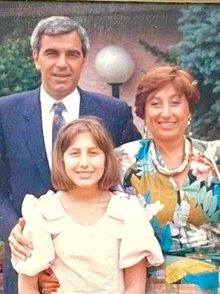 A man, woman, and young girl posing outdoors, smiling. The man is wearing a dark suit and tie, the woman is in a colorful blouse and necklace, and the girl is in a white dress.