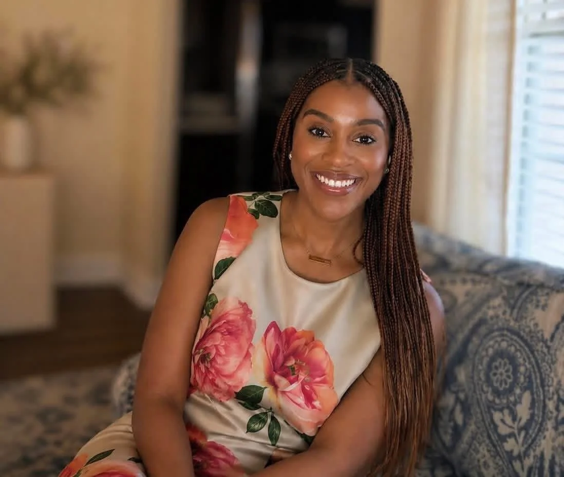 A smiling woman with braids sitting on a patterned couch in a cozy room, wearing a floral sleeveless dress, with sunlight coming through a window.