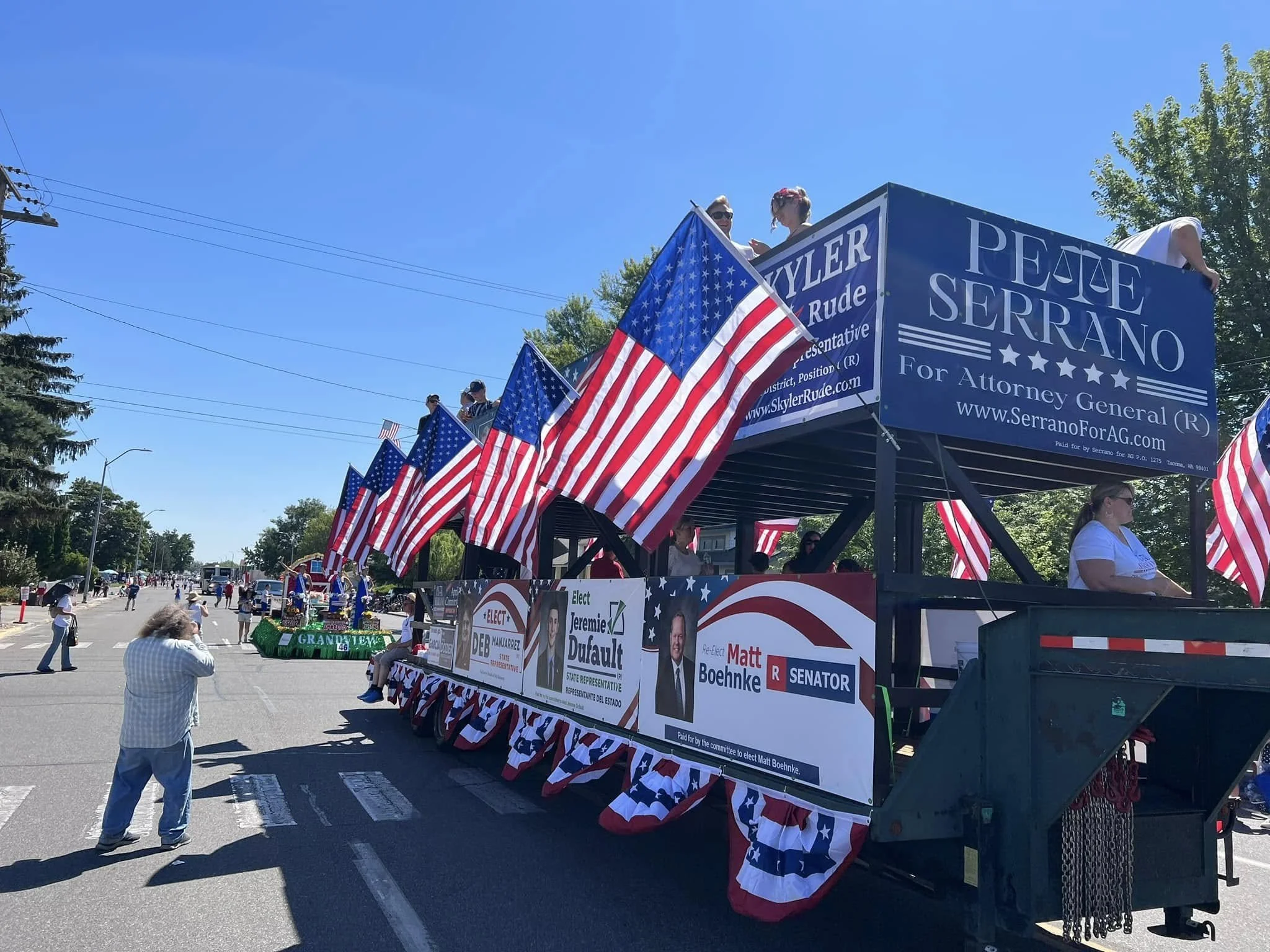 A parade float decorated with American flags, promoting political candidates, with onlookers watching from the sidewalk on a sunny day.
