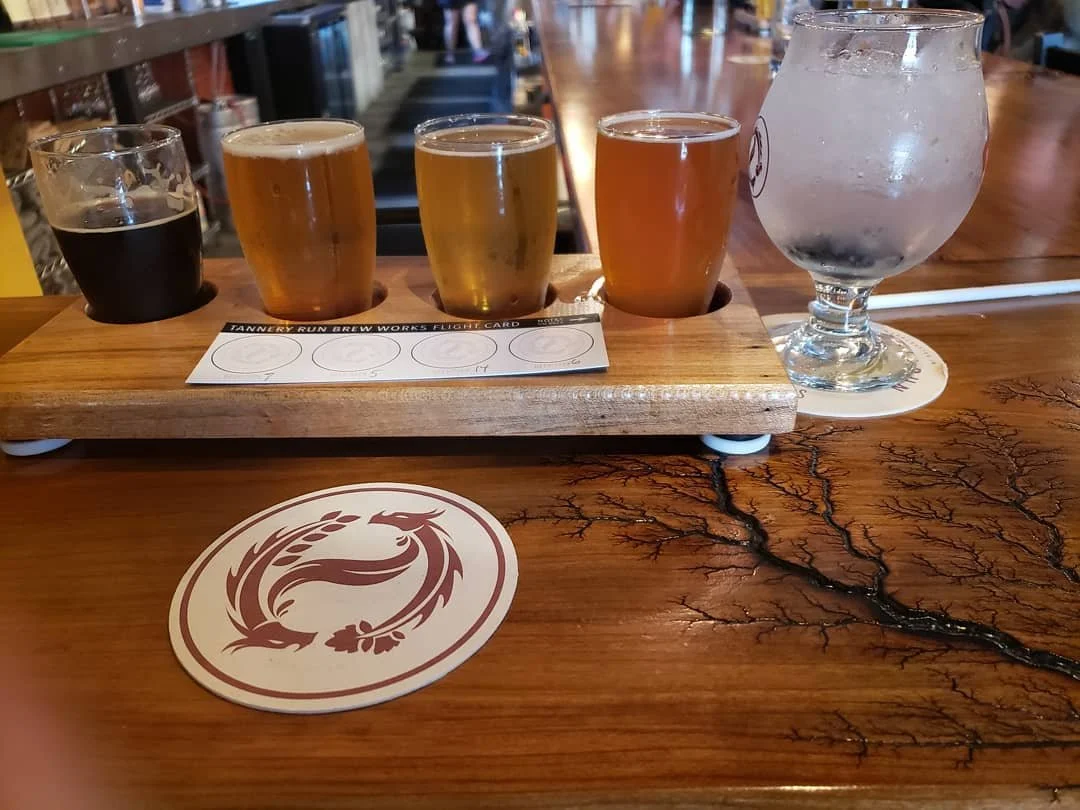 A flight of four small glasses of different beers on a wooden paddle, accompanied by a large glass of water on a coaster, placed on a wooden bar counter.
