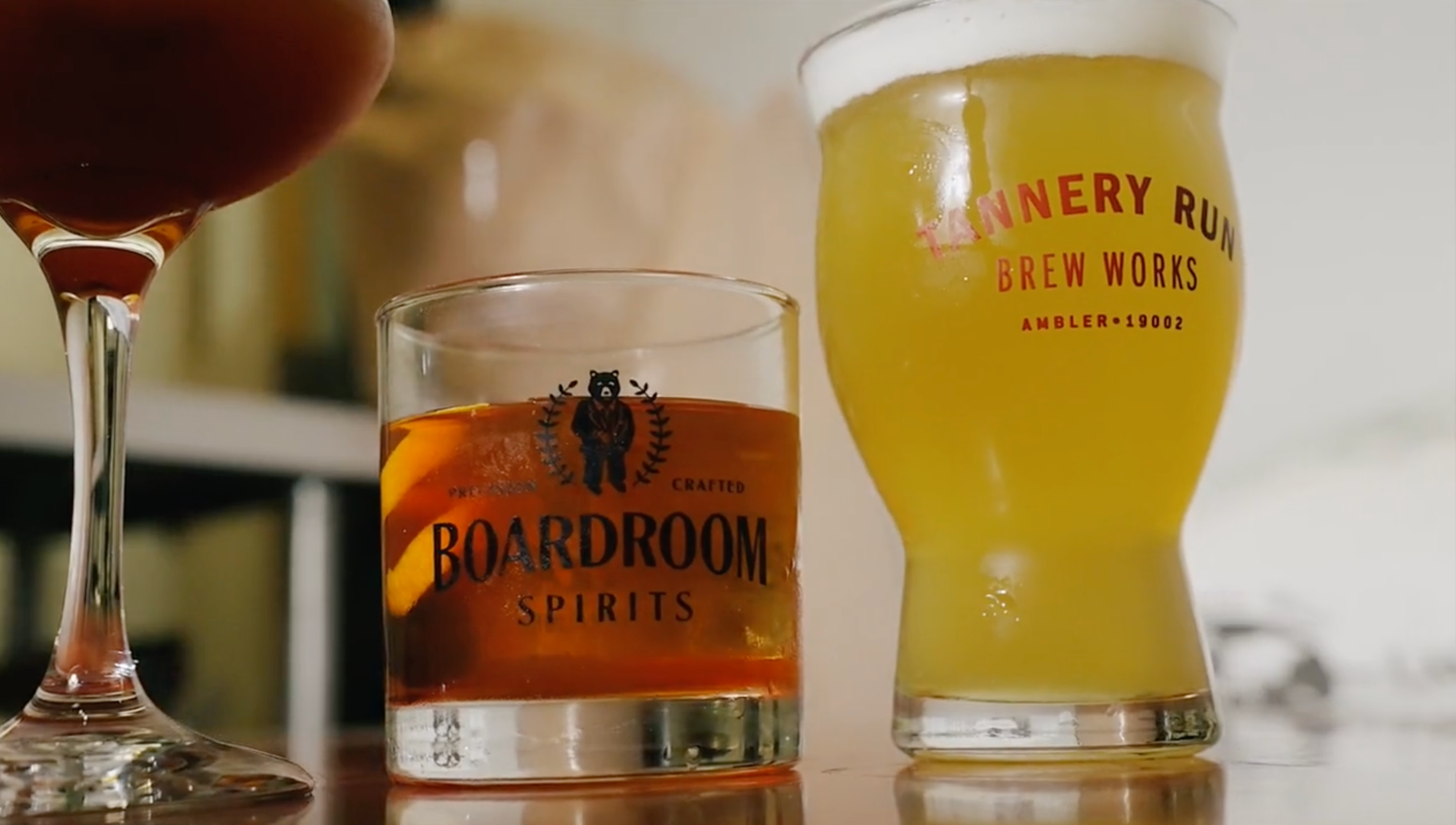 Close-up of three beverages on a wooden surface: a dark red wine in a flute glass, an orange-hued spirit in a glass labeled 'Boardroom Spirits' with lemon slices, and a yellow cocktail in a tall glass with red text 'Tannery Run Brew Works'.
