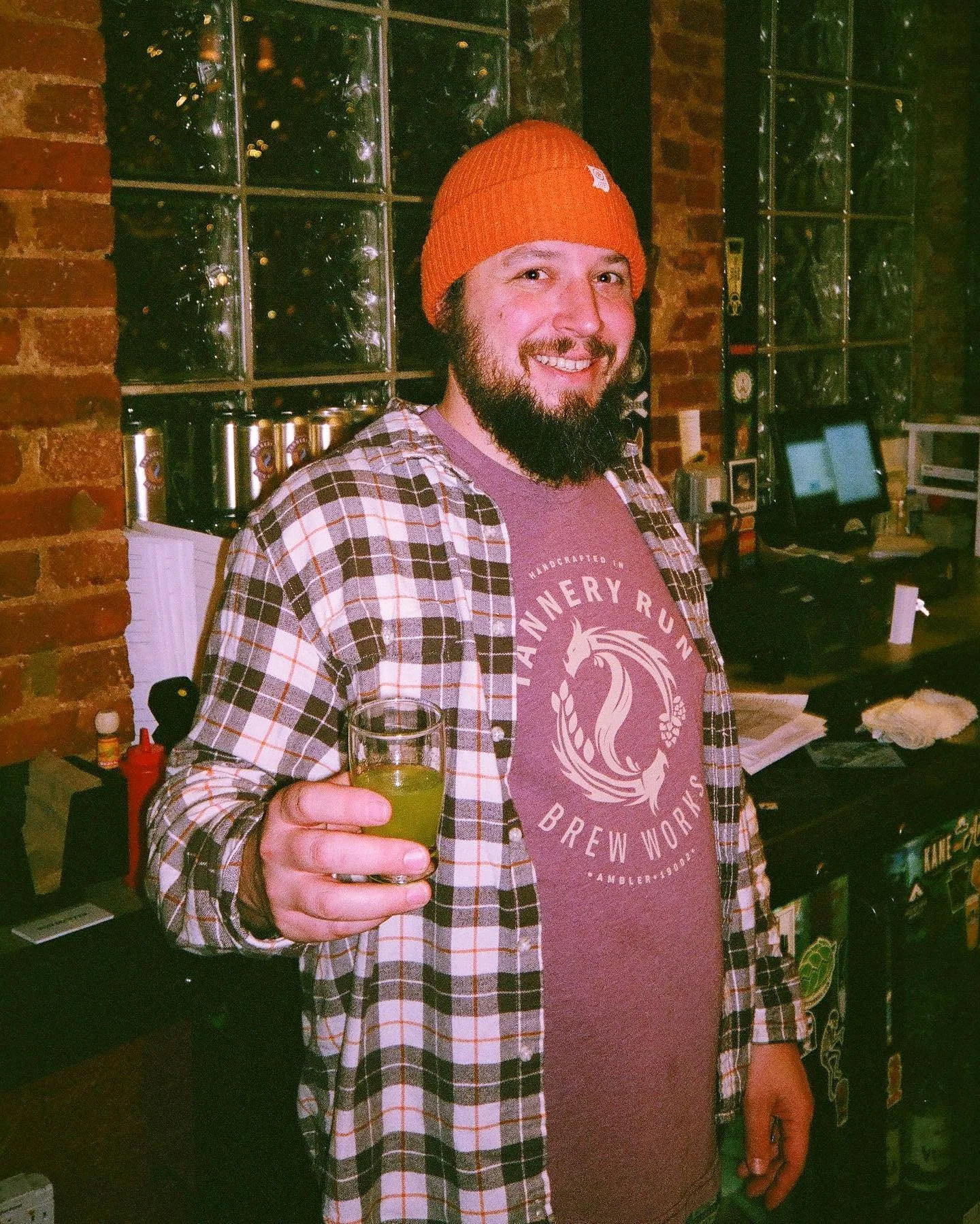 A man with a beard and mustache, wearing an orange knit beanie and a plaid shirt over a pink T-shirt, is smiling and holding a glass of  in his right hand. He is standing indoors near a brick wall and behind him are shelves with bottles, a computer monitor, and other items.