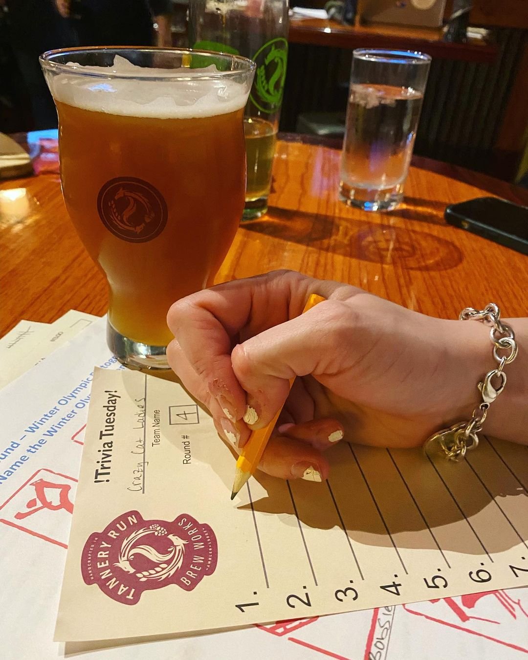 A person with a silver bracelet and painted nails holding a pencil, writing on a quiz sheet at a wooden table. Three beverages are on the table: an amber beer in a glass, a green bottle of beer, and a glass of water.