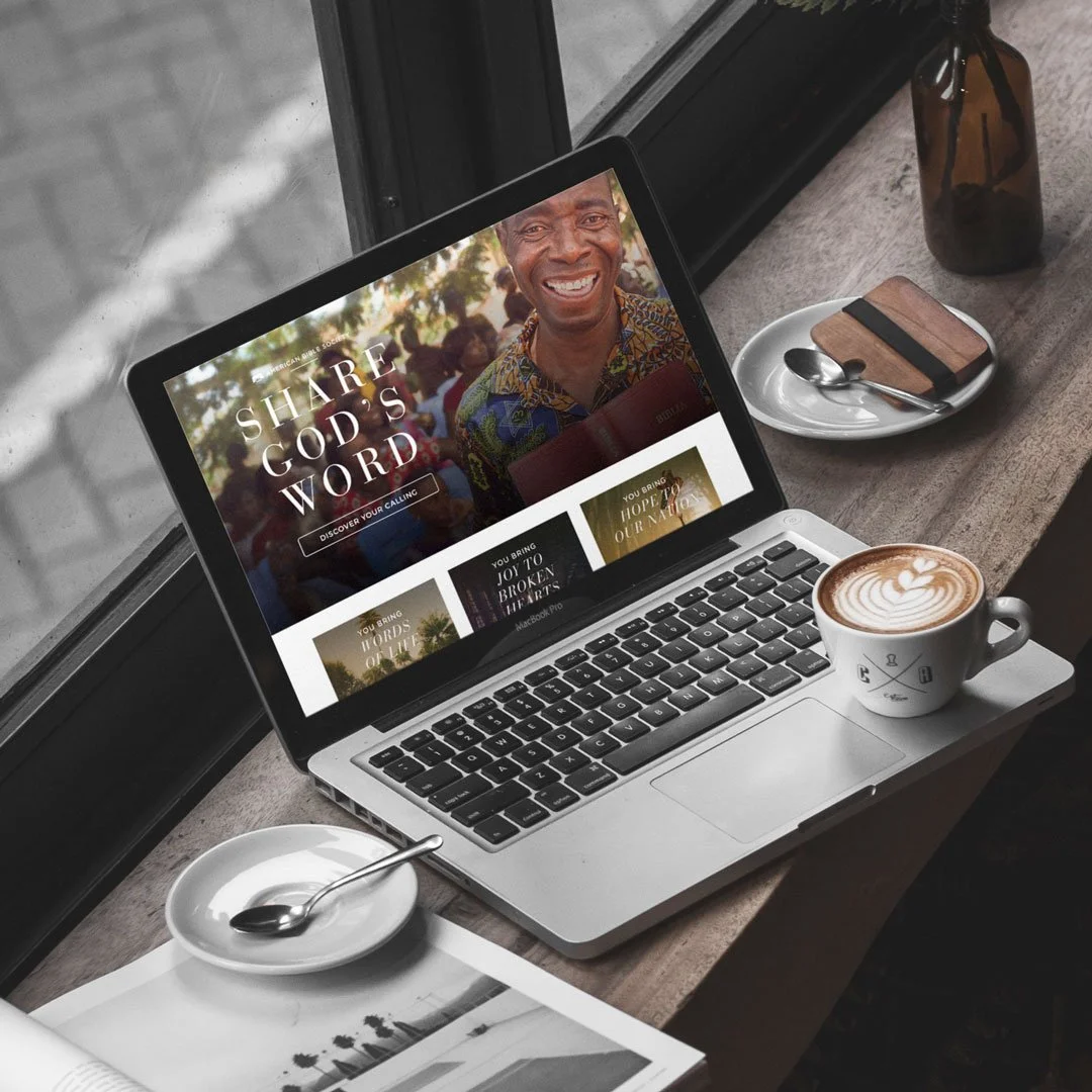 A laptop displaying a website about sharing God's word, placed on a wooden table near a window. Next to the laptop are two small plates with spoons, a coffee cup with latte art, a small brown bottle, and a closed notebook with a spoon on top of it.