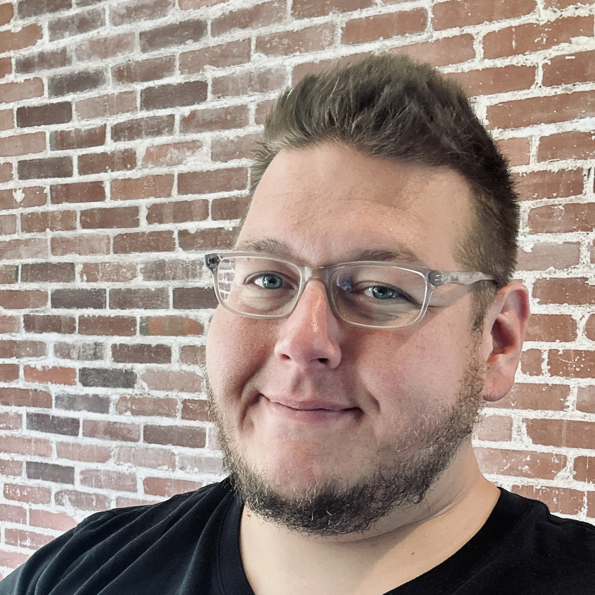 Close-up of a man with short brown hair, glasses, and facial hair, wearing a black shirt, standing in front of a brick wall.