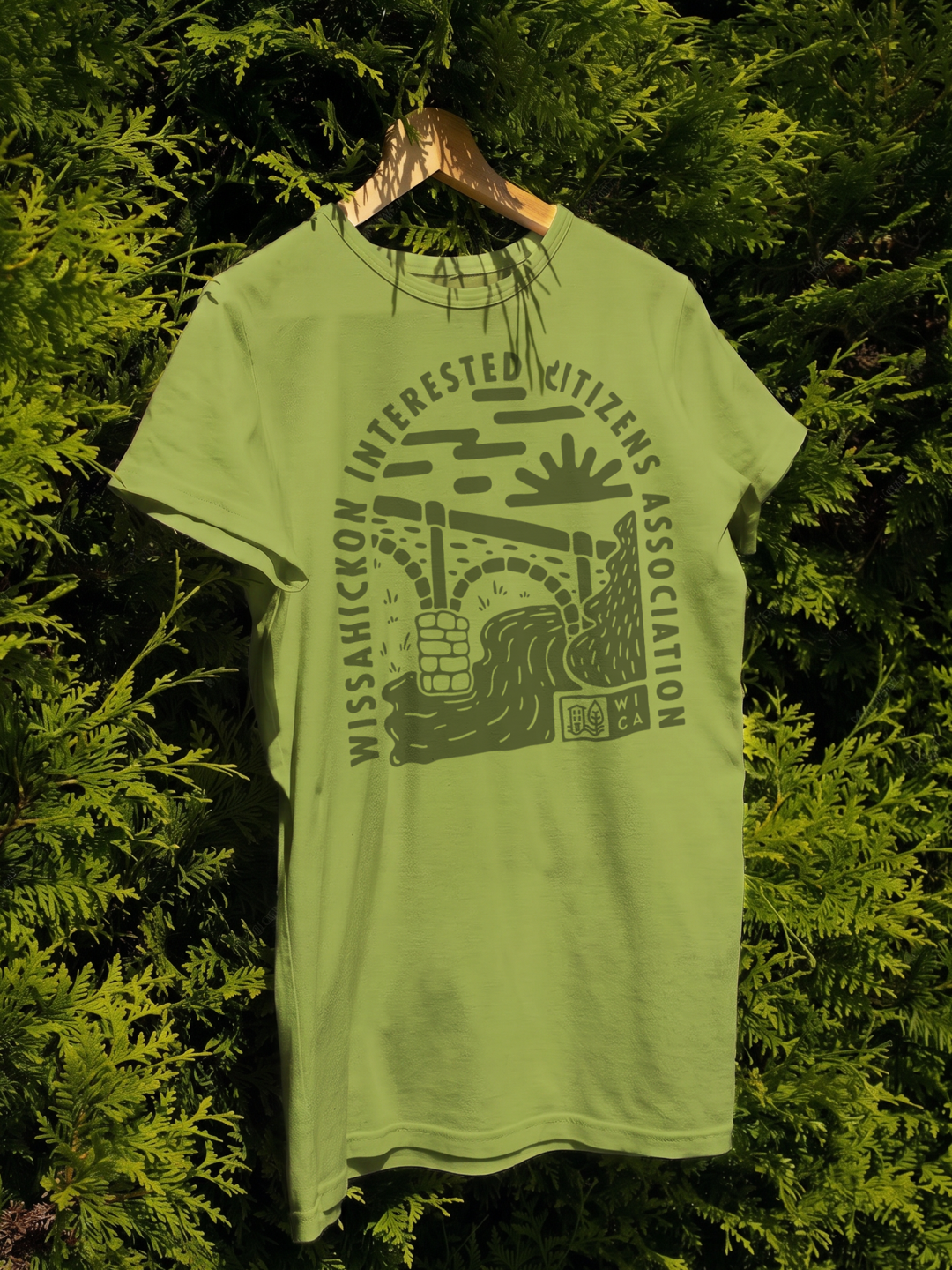 Green T-shirt with black graphic design hanging on a wooden hanger in front of lush greenery. The design features a bridge, arrow, water, and a sign that reads 'WCA'. Circular text says 'Missahickon Interested Citizens Association'.