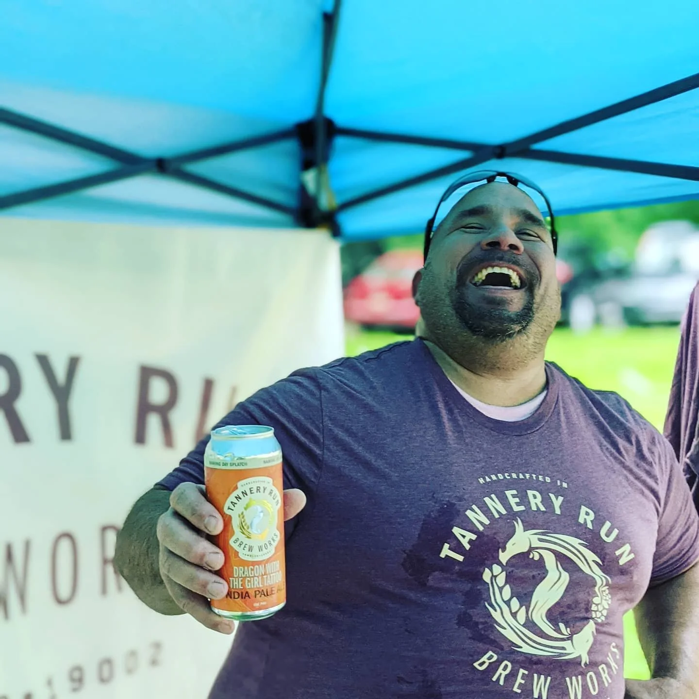 A man with glasses and a beard smiling and holding a can of beer at an outdoor event under a blue canopy.