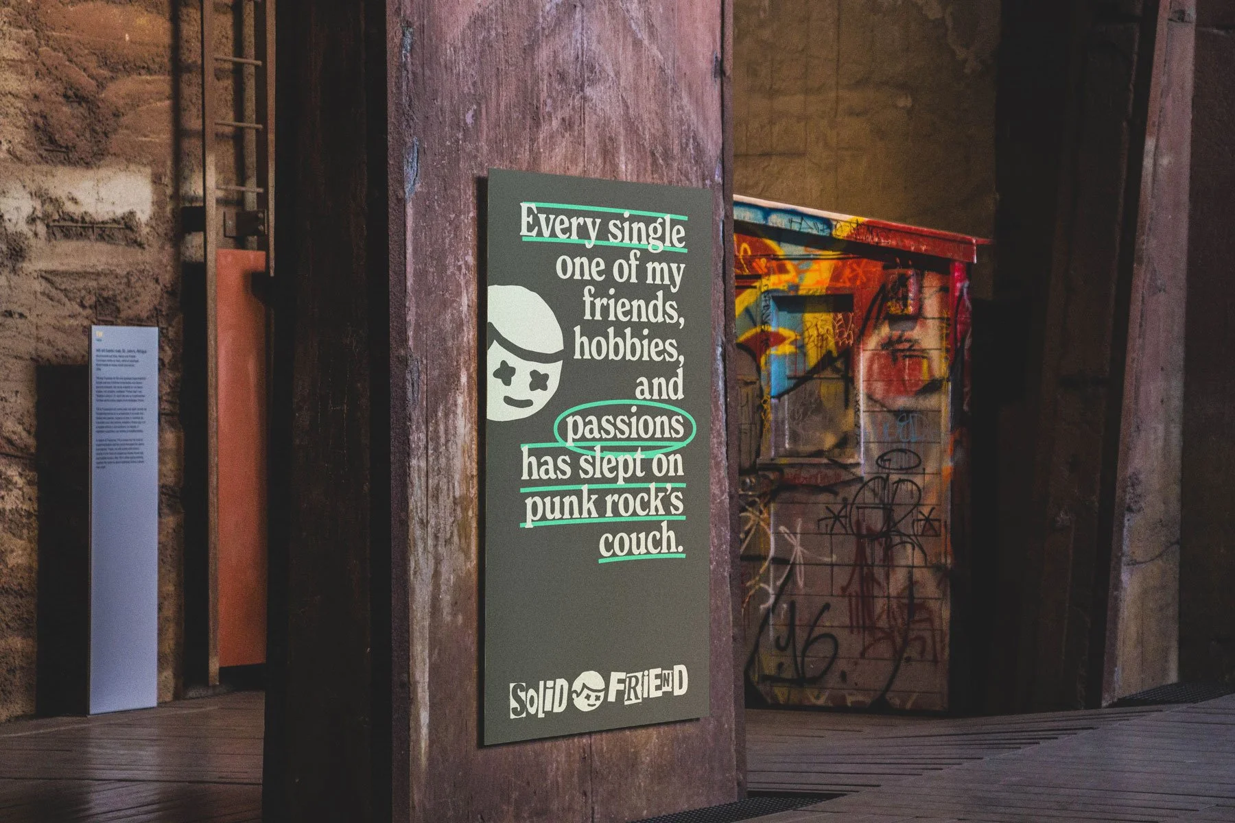 A sign on a wall reading: 'Every single one of my friends, hobbies, and passions has slept on punk rock's couch.' The wall behind has graffiti art, and there is a wooden floor and other walls with stone and graffiti designs.