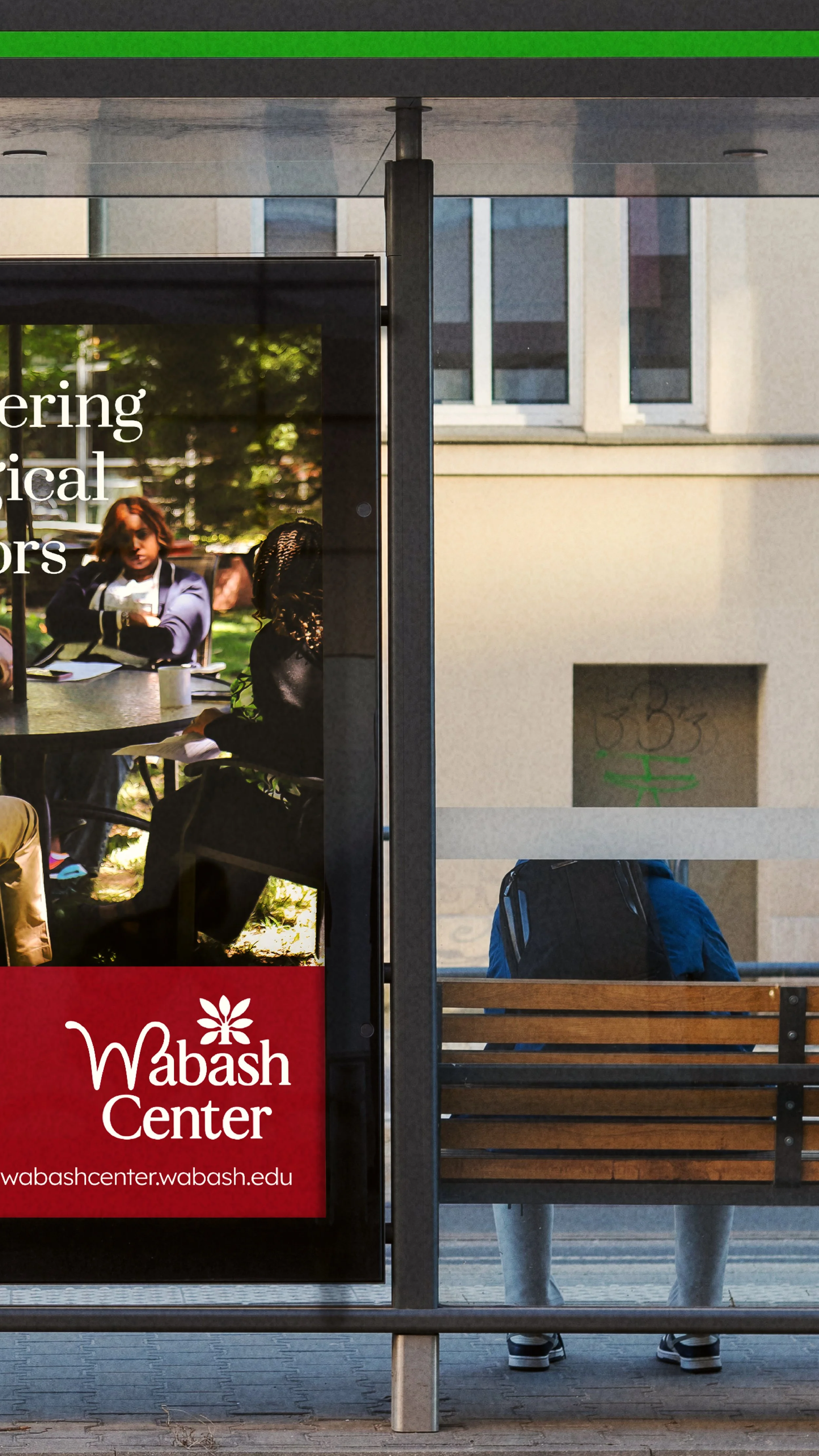 A person with a blue backpack sitting on a wooden bench at a bus stop, with a large sign displaying the Wabash Center logo and website, and part of a building in the background.