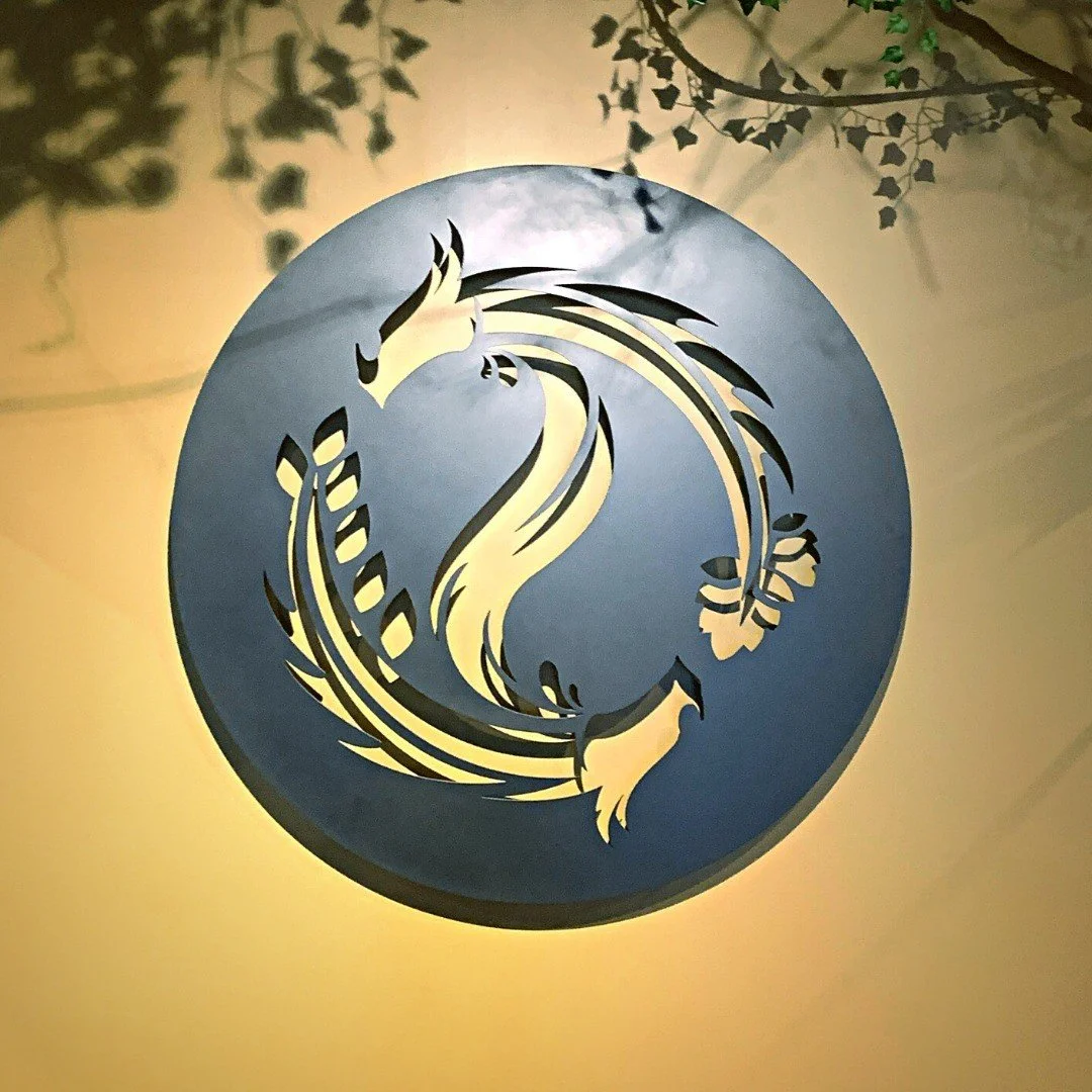 A circular decorative wall piece with a stylized split dragon design, in gold and black. Shadows of leaves and branches cast on the wall behind it.