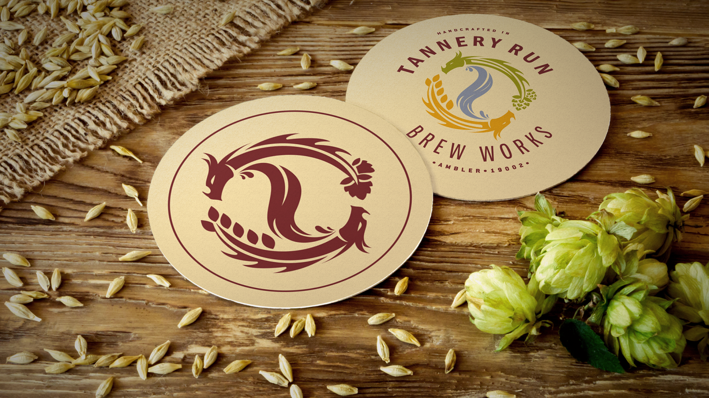 Two circular bar coasters for Tannery Run Brew Works on a wooden surface, one featuring a dragon design and the other displaying the brewery's logo with a flowing water image and hops, with scattered barley grains and green hop flowers around.