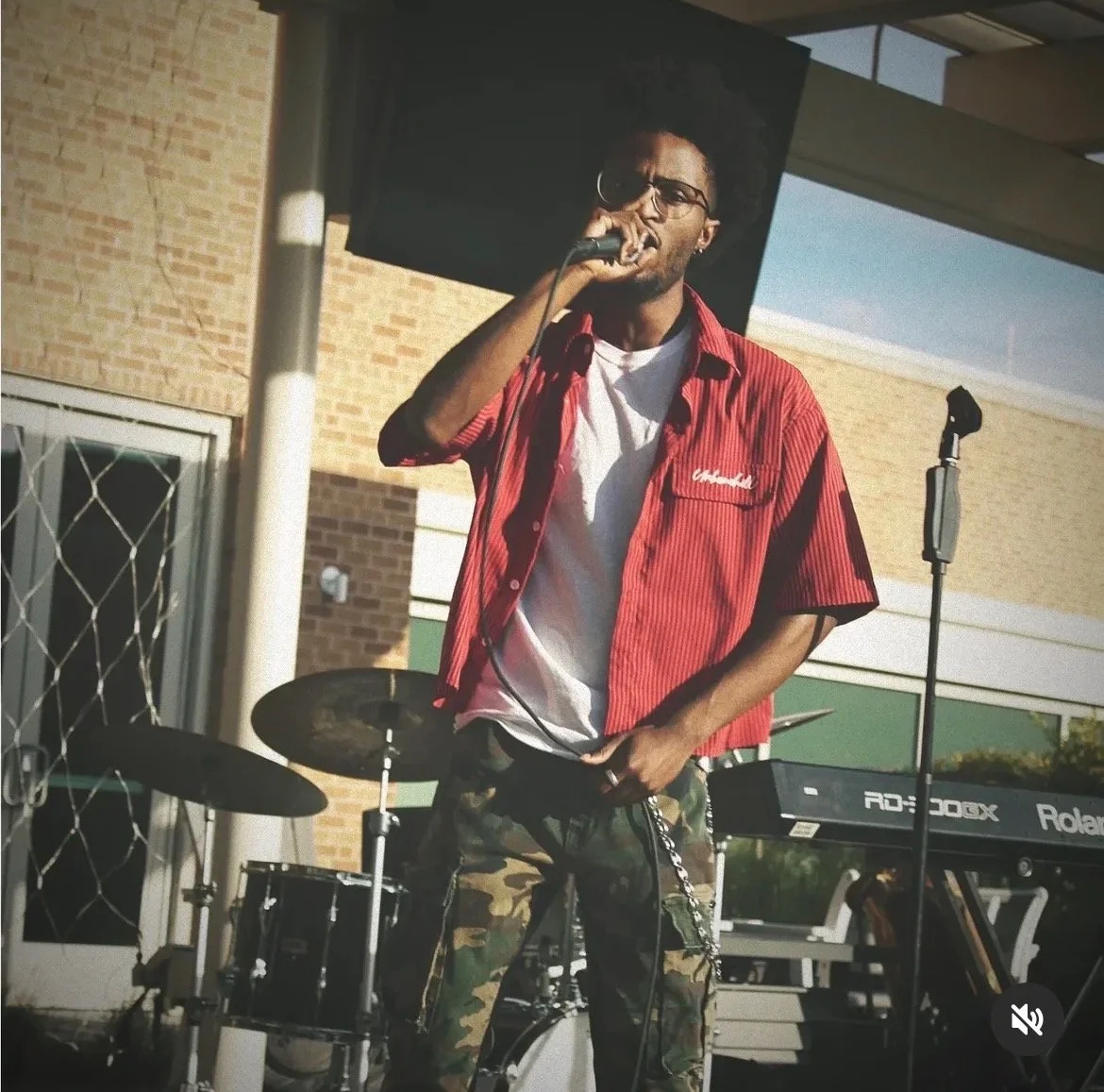 A young man singing into a microphone outdoors, standing near a drum set and keyboard on a stage.